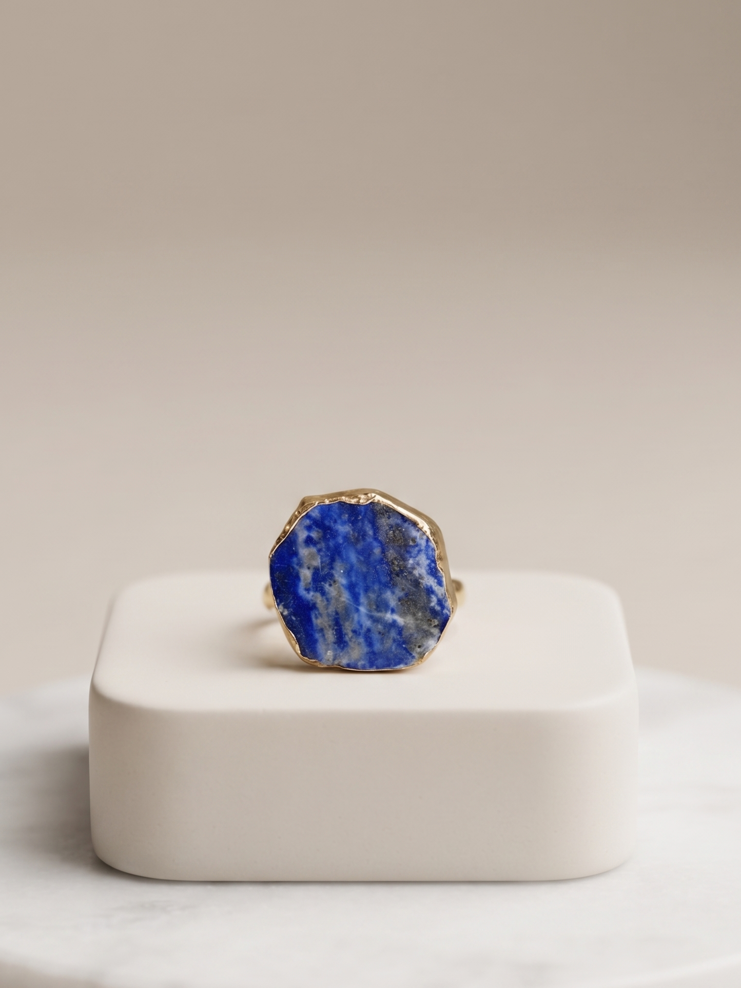 A gold ring with a large, irregular-shaped blue lapis lazuli stone, displayed on a white square pedestal over a round white marble tray against a warm beige background.