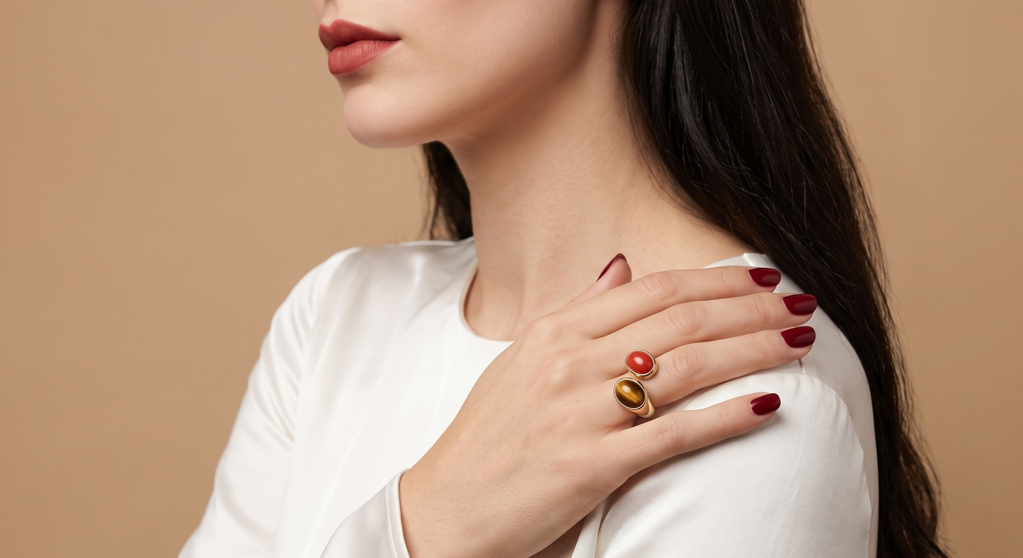 A close-up lifestyle shot of a woman wearing a gold ring with a red stone on her finger, dressed in a white top against a solid beige background.
