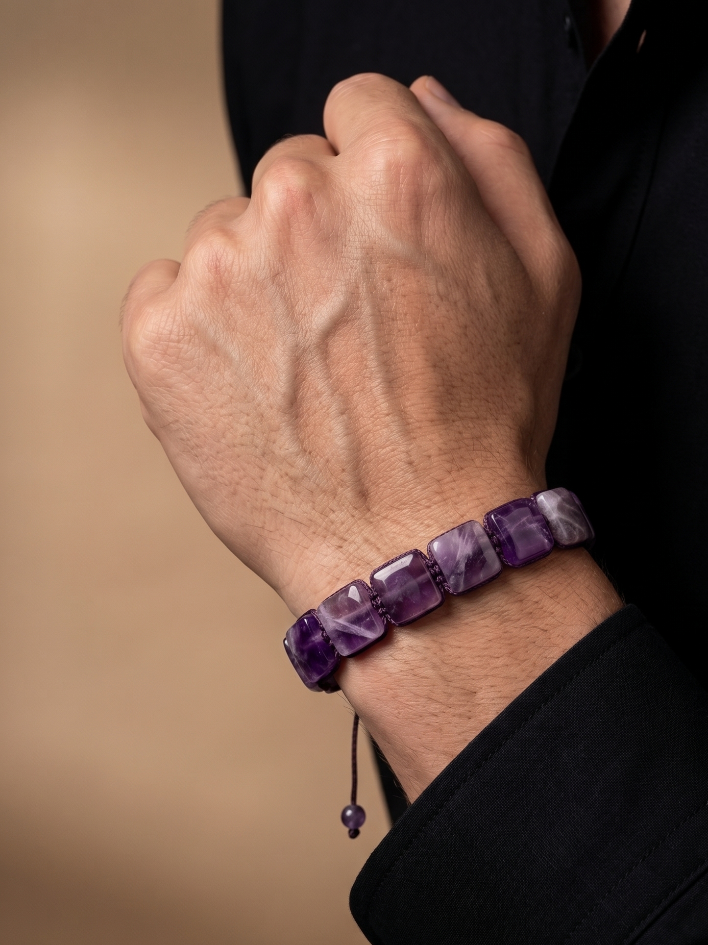 A close-up of a person's hand wearing a purple amethyst beaded bracelet, dressed in a black shirt against a warm beige background.
