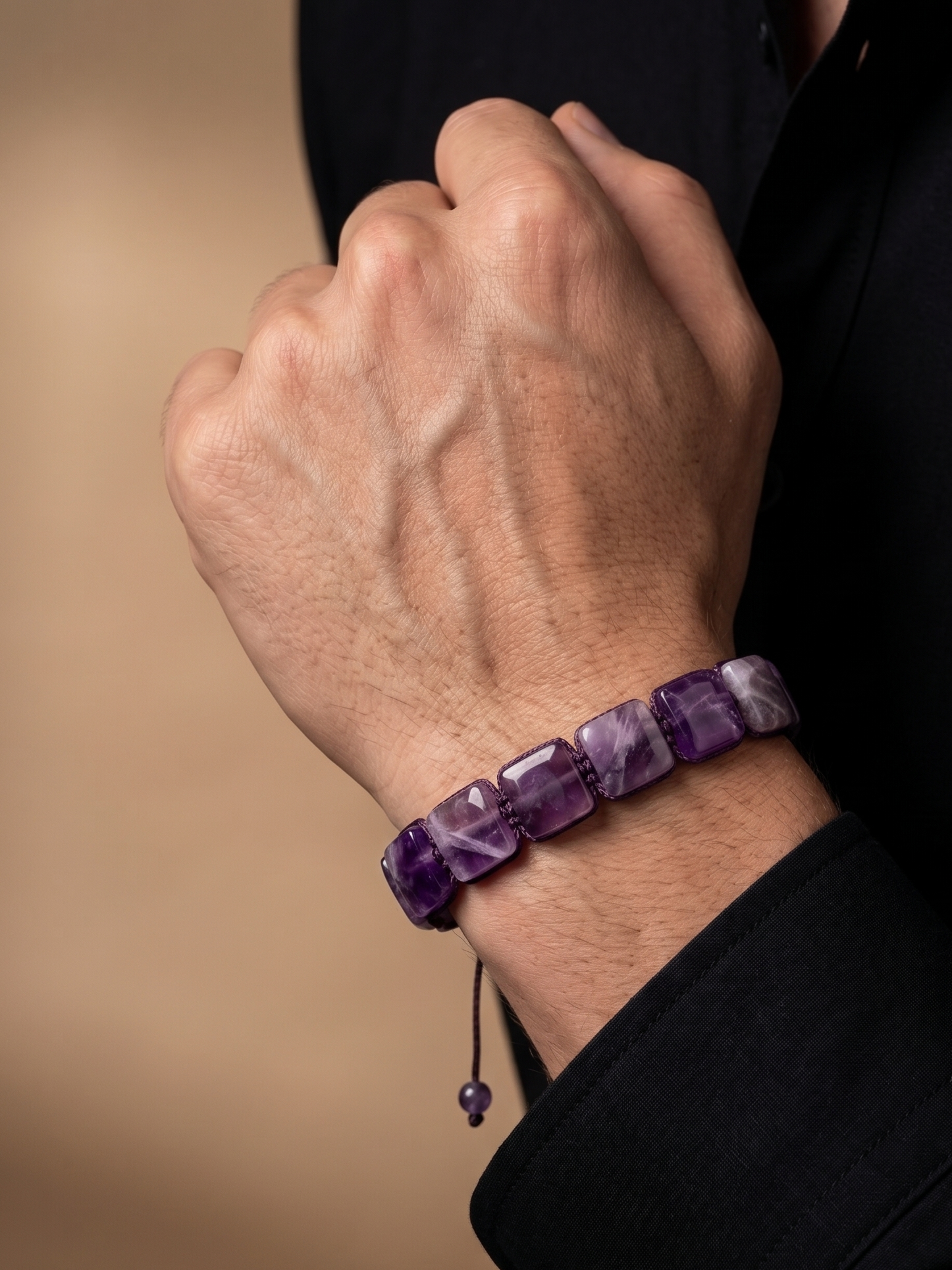 A close-up of a person's hand wearing a purple amethyst beaded bracelet, dressed in a black shirt against a warm beige background.