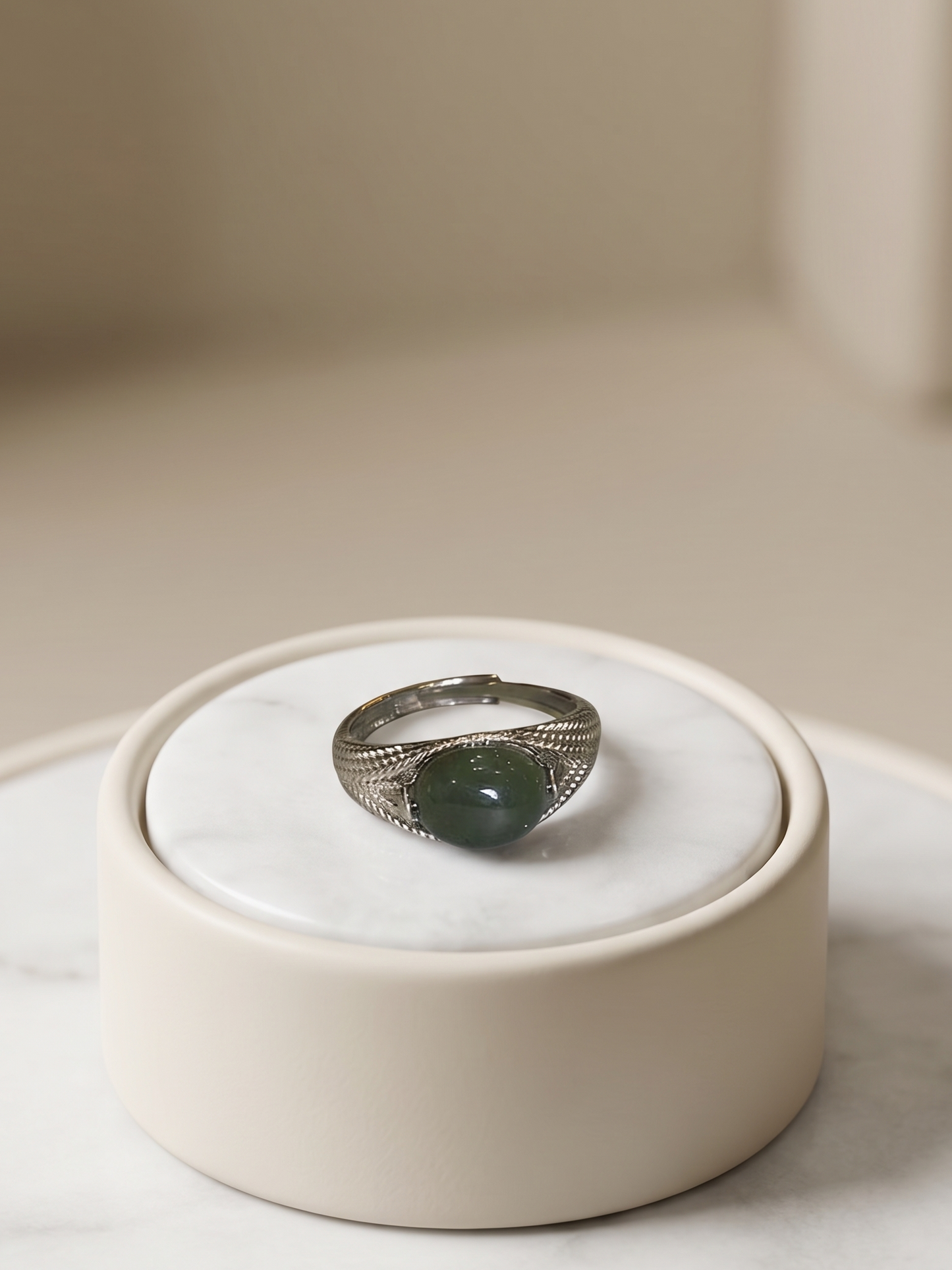 A silver ring with a dark green oval stone, displayed on a small round white marble stand.