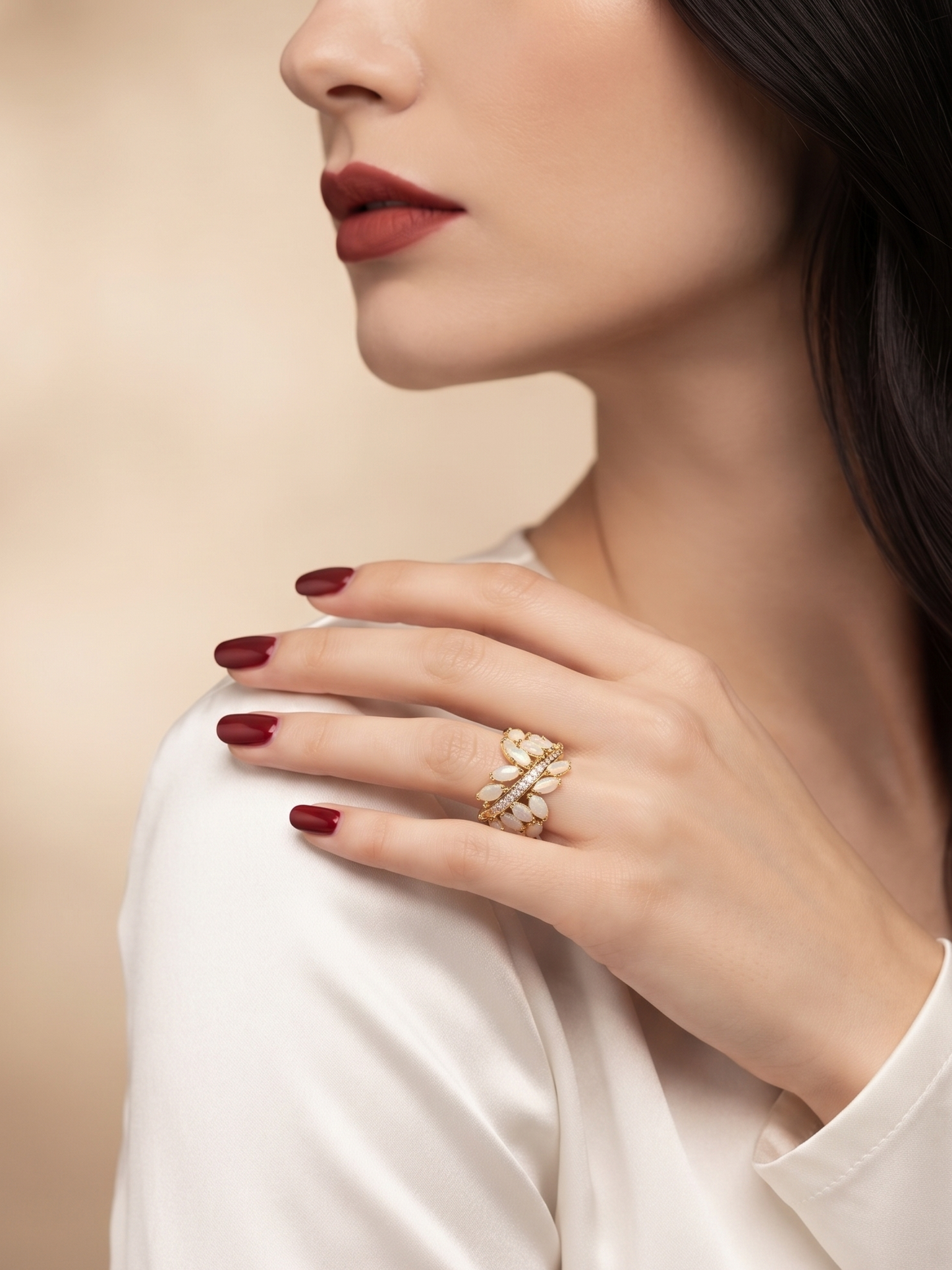 A close-up lifestyle shot of a woman wearing a gold ring with a floral or butterfly-like design on her finger, paired with a white satin top and dark red nail polish against a soft beige background.