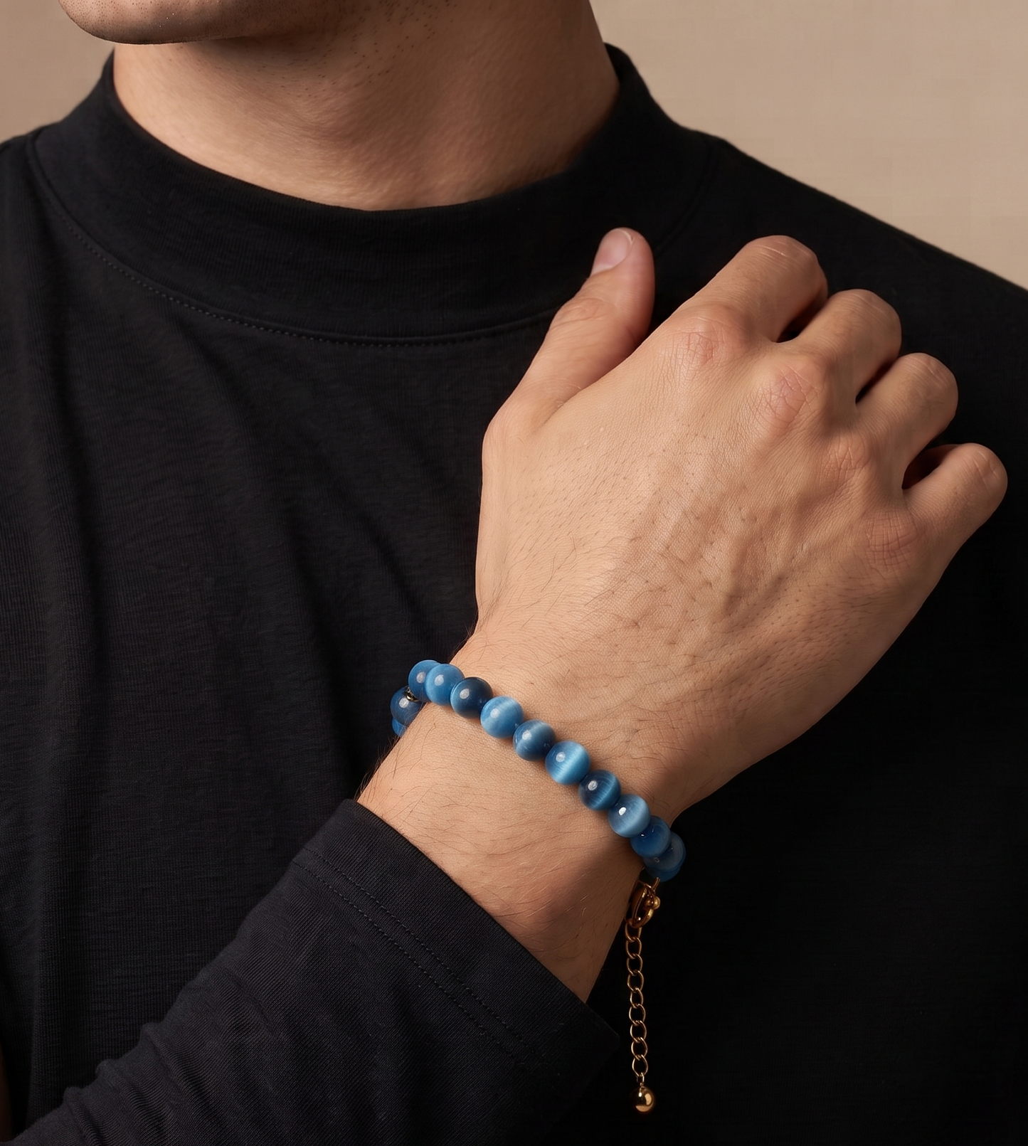 A close-up of a man wearing a dark navy blue beaded bracelet with a gold chain extender on his wrist, paired with a black long-sleeve shirt against a warm beige background.
