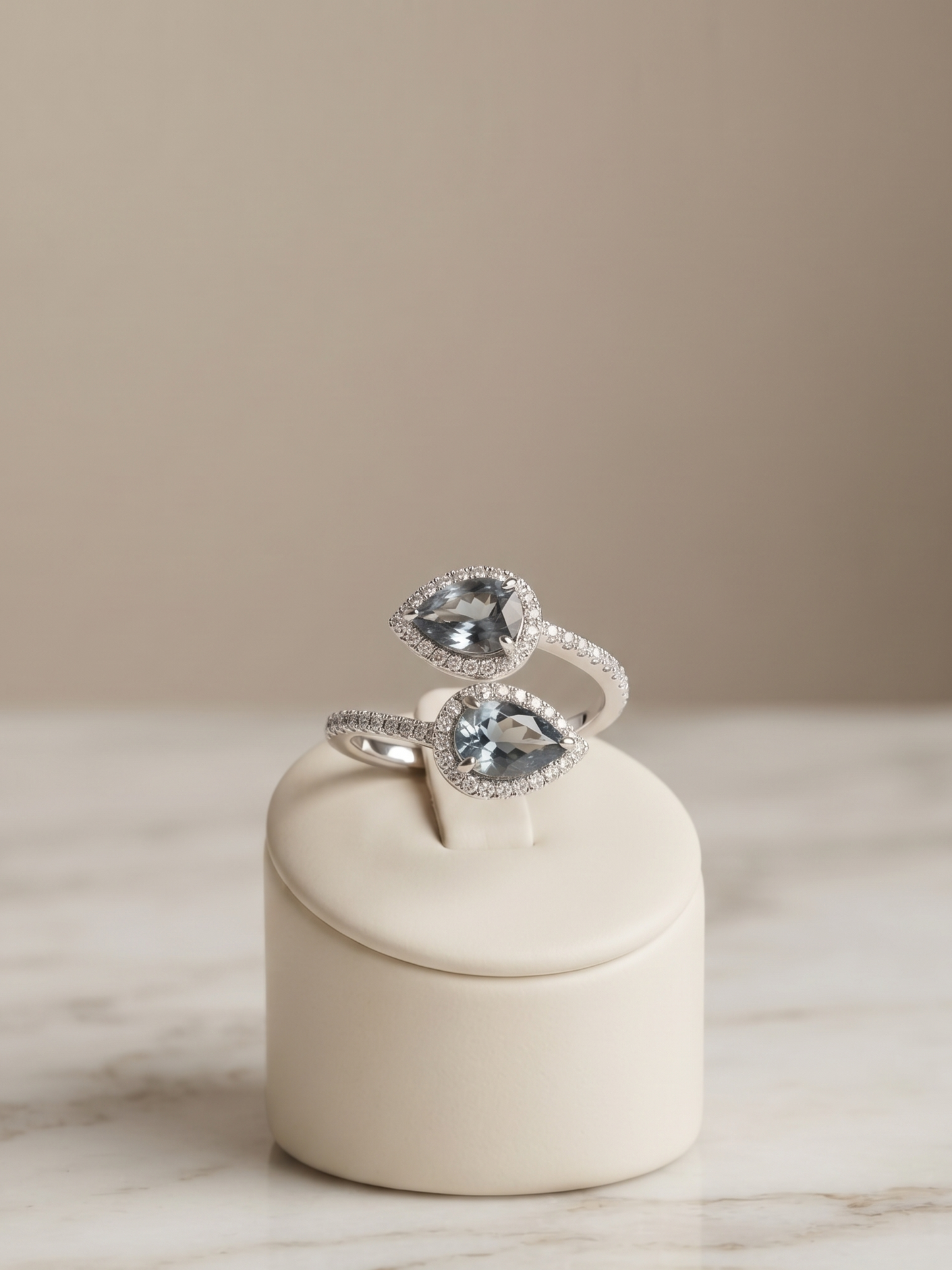 A silver ring with two pear-cut clear gemstones, each surrounded by small pavé-set clear stones, displayed on a cream cylindrical ring stand over a marble surface against a warm beige background.