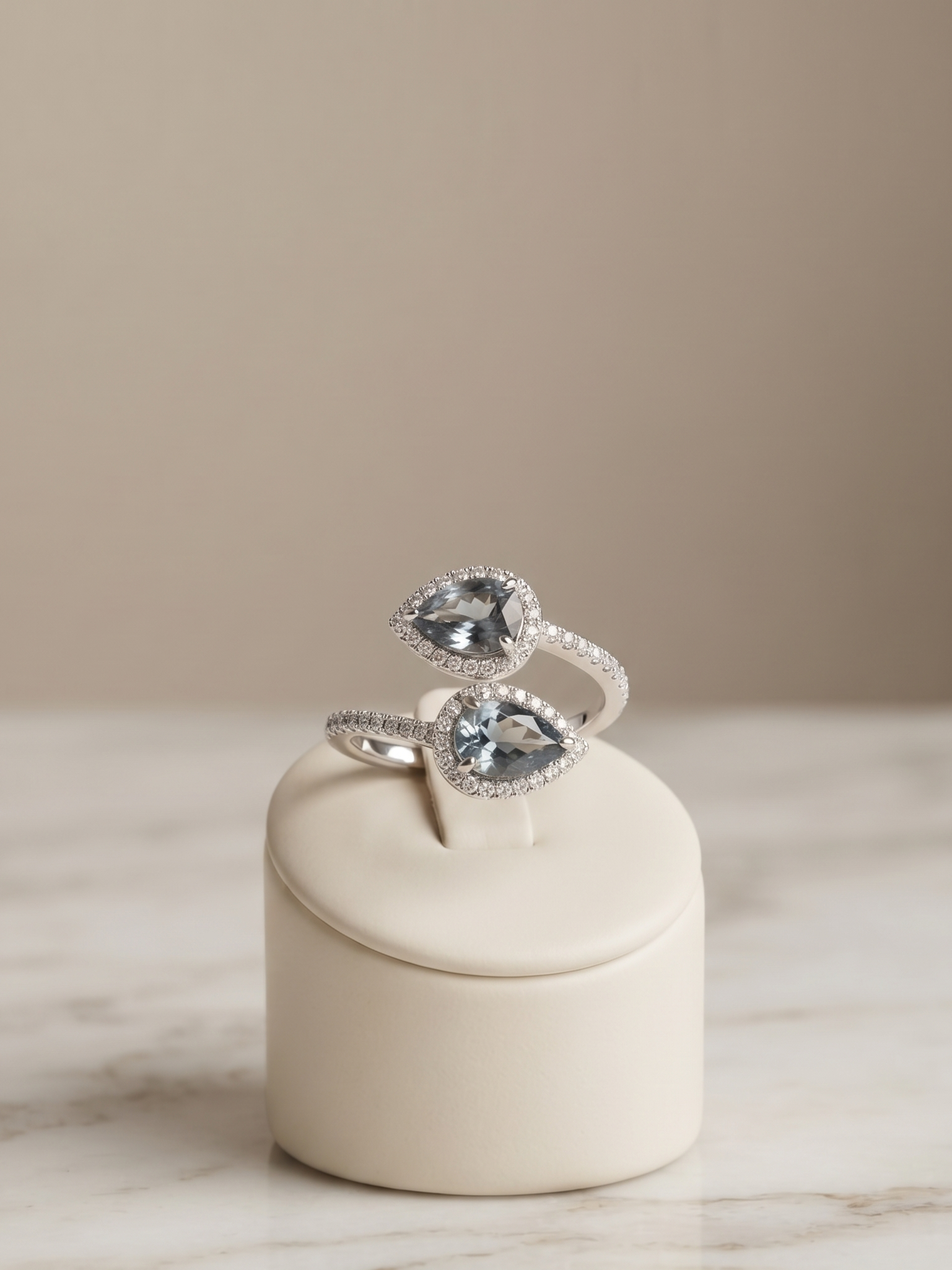A silver ring with two pear-cut clear gemstones, each surrounded by small pavé-set clear stones, displayed on a cream cylindrical ring stand over a marble surface against a warm beige background.