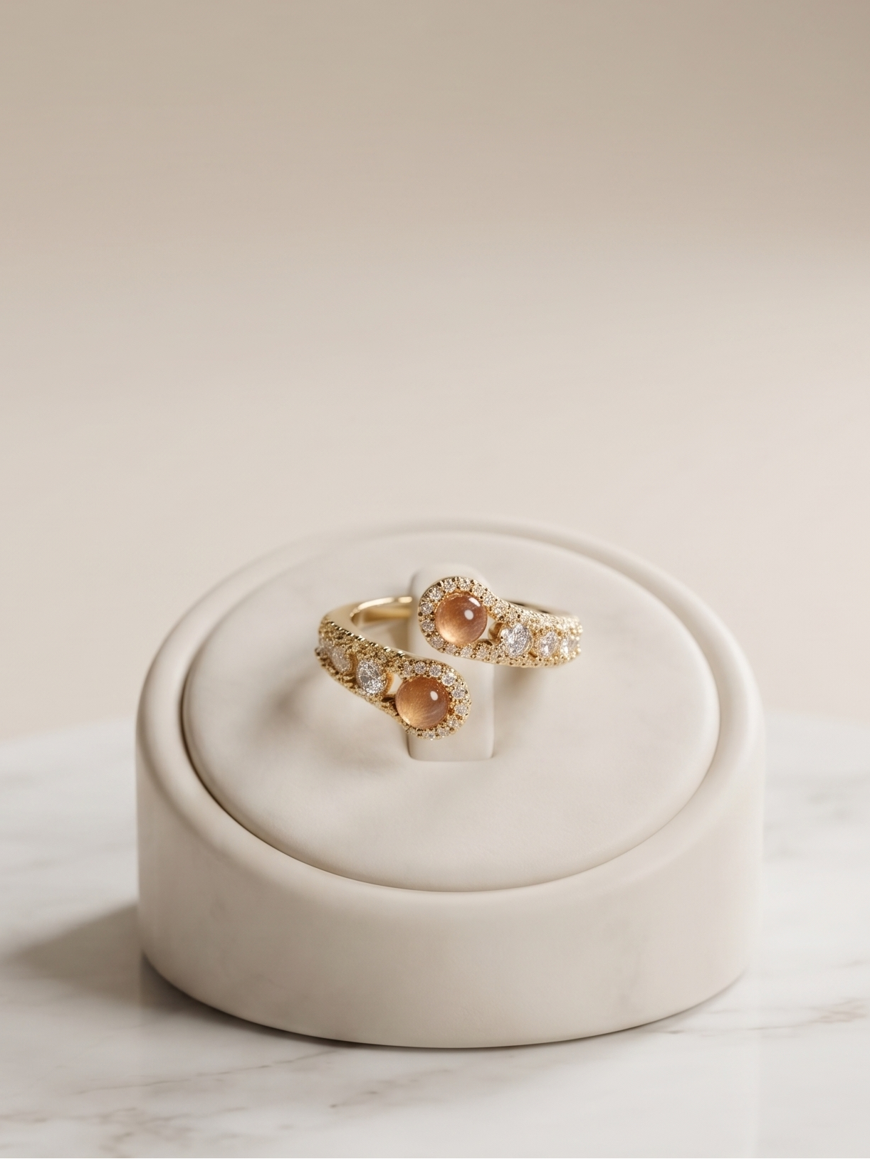 Two gold rings with oval peach-orange center stones and small clear accent stones, displayed on a round white jewelry stand atop a white marble surface against a warm beige background.