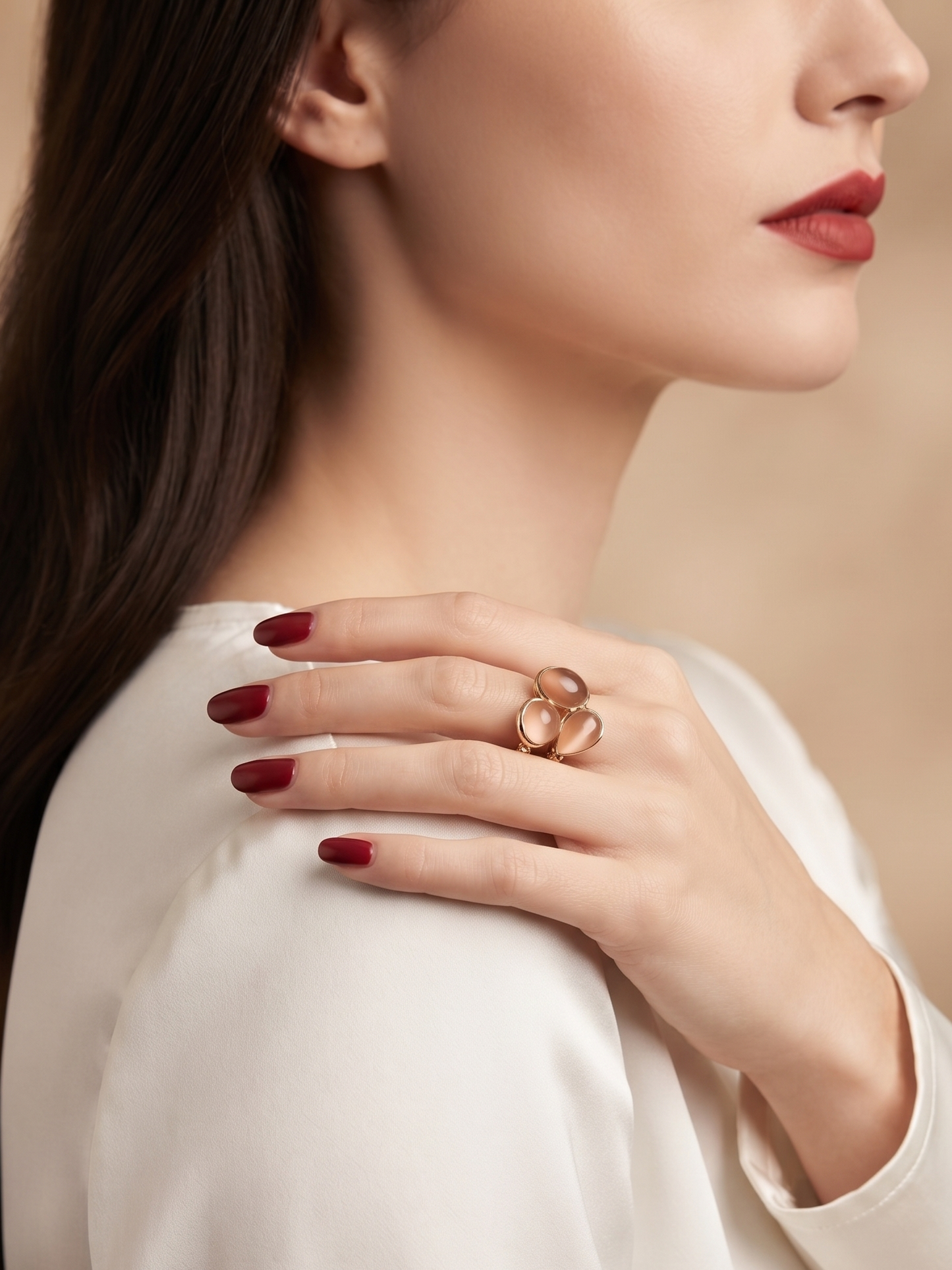 A close-up lifestyle shot of a woman wearing a rose gold double-ring ring on her hand, which is resting on her shoulder. She is wearing a white satin blouse and has dark red manicured nails, set against a soft, warm beige background.