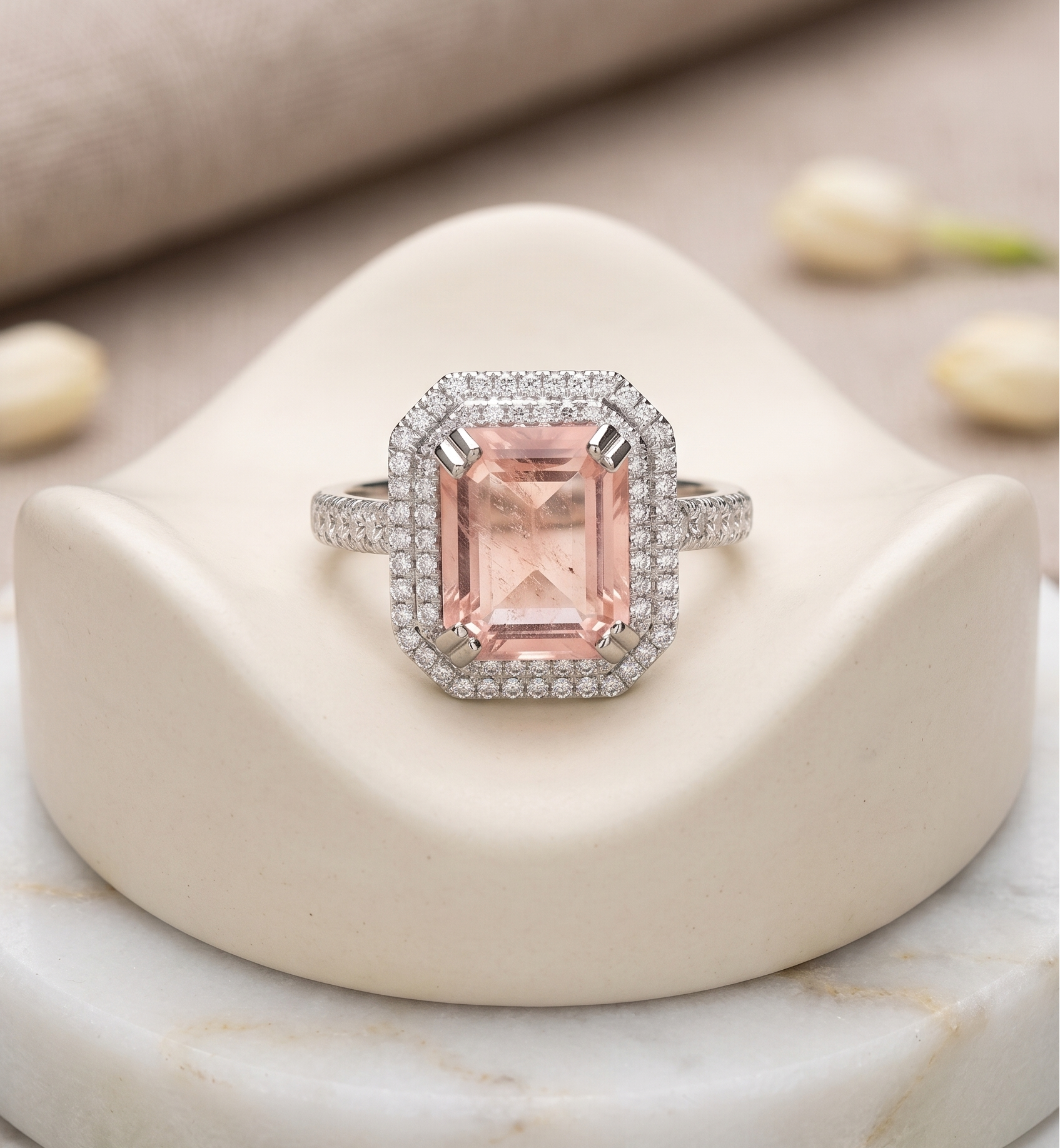 A halo ring featuring a rectangular emerald-cut pink gemstone surrounded by a silver-tone halo and pavé setting, displayed on a cream-colored jewelry stand.
