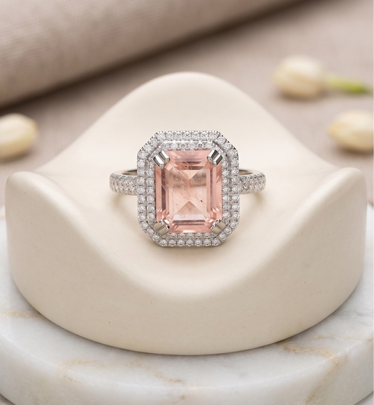 A halo ring featuring a rectangular emerald-cut pink gemstone surrounded by a silver-tone halo and pavé setting, displayed on a cream-colored jewelry stand.