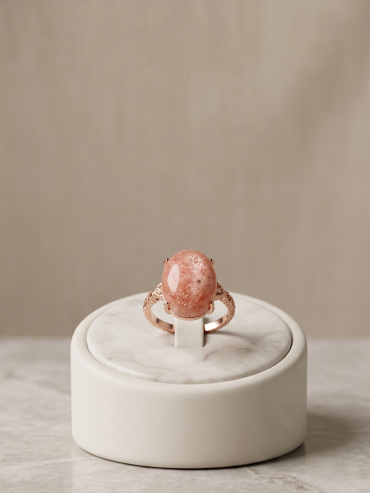 A rose gold ring with a large, oval, pinkish-orange stone set on a small white marble display stand, placed on a light marble surface against a soft beige background.