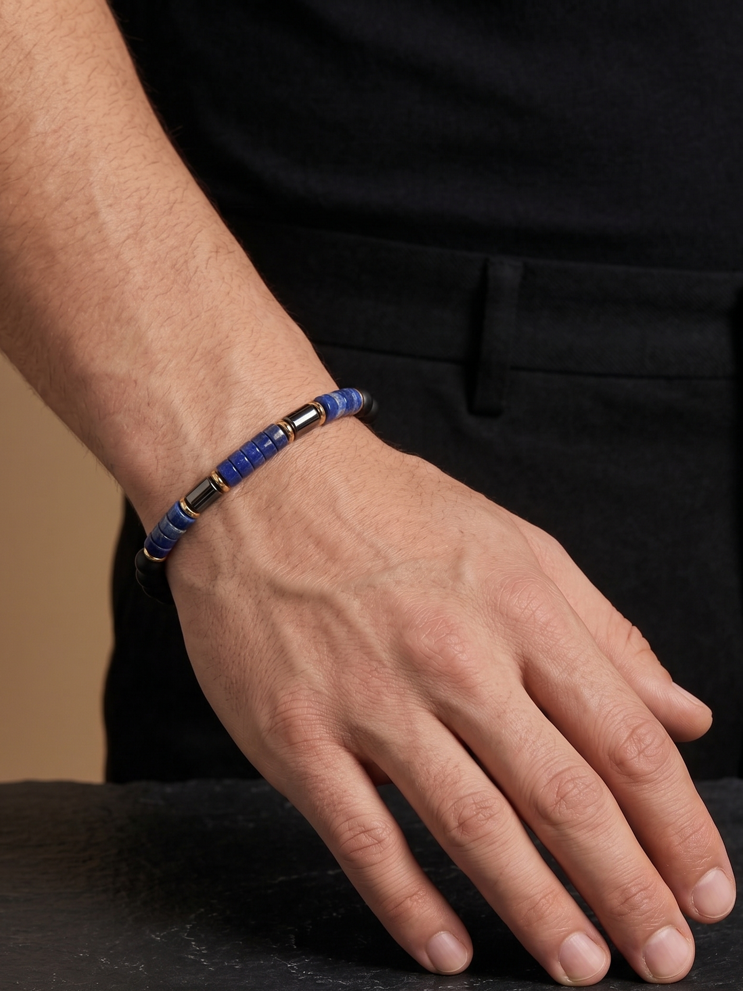 A close-up of a person's hand wearing a beaded bracelet with blue, black, and metallic gold-toned beads, resting on a dark slate surface.