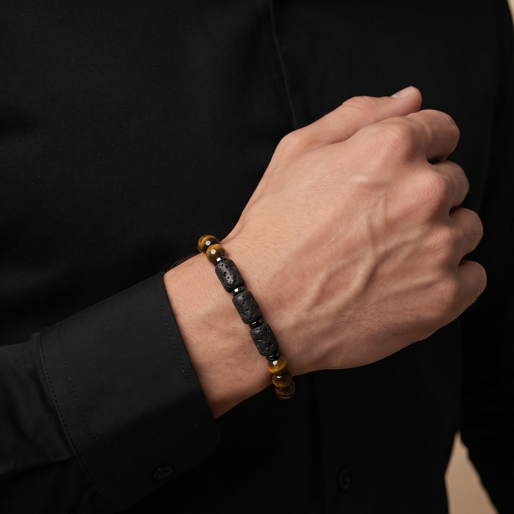 A close-up of a man's hand and wrist wearing a beaded bracelet. The bracelet features dark gray polished stone beads, a small gold spacer bead, and two tiger's eye beads at the ends.