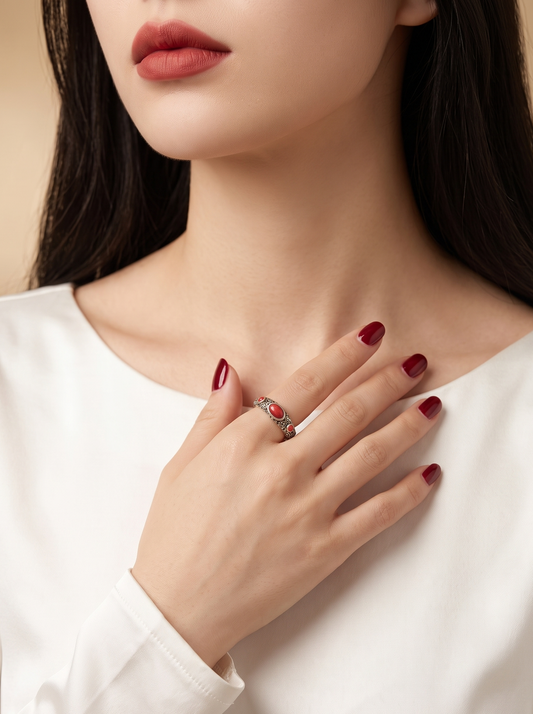 A close-up of a woman wearing a silver ring with a red heart-shaped stone on her ring finger, paired with a white long-sleeve top and dark red manicured nails.
