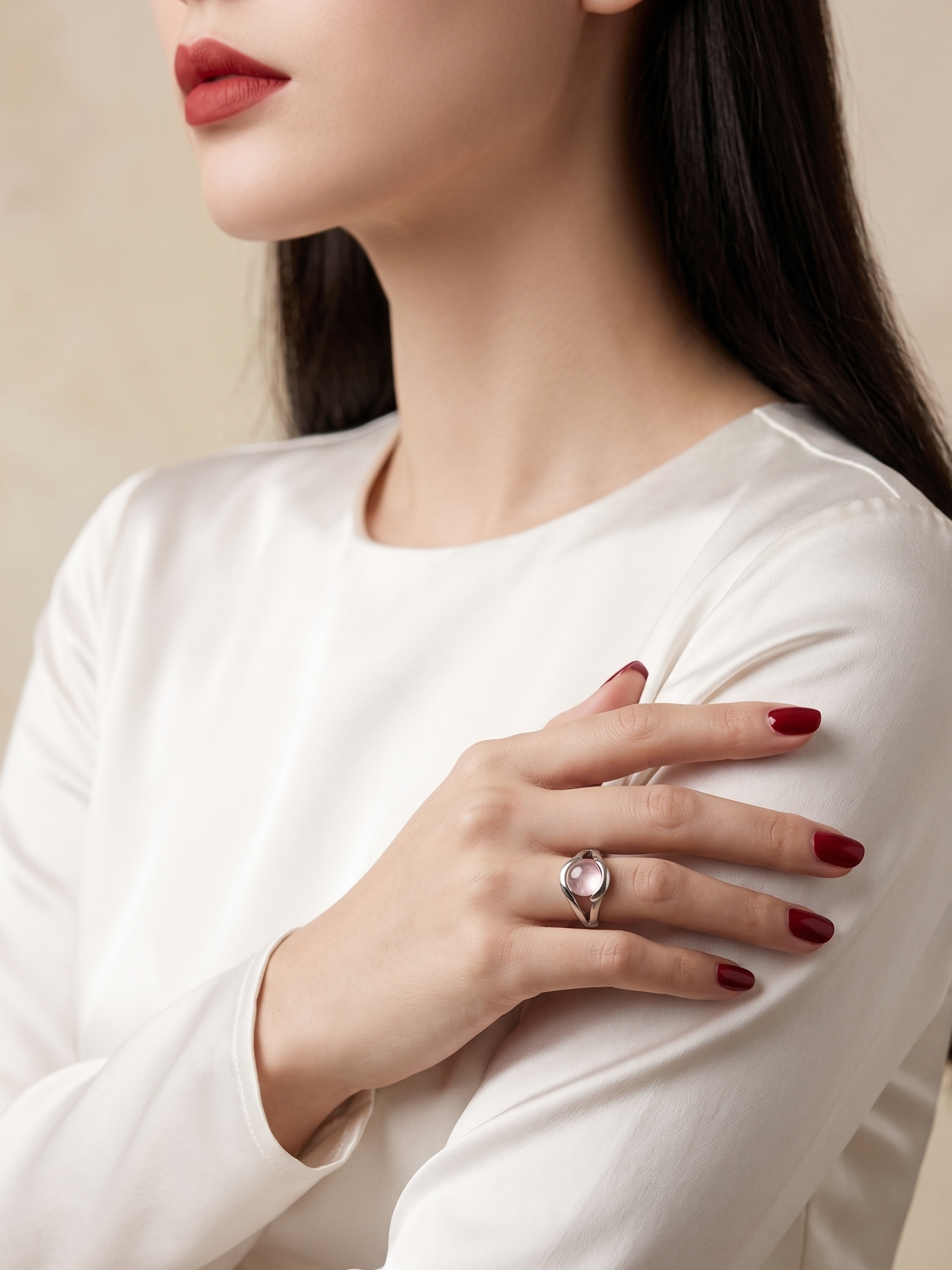 A close-up of a woman wearing a silver ring with a clear oval gemstone on her finger, paired with a white long-sleeve top and dark red nail polish against a soft beige background.