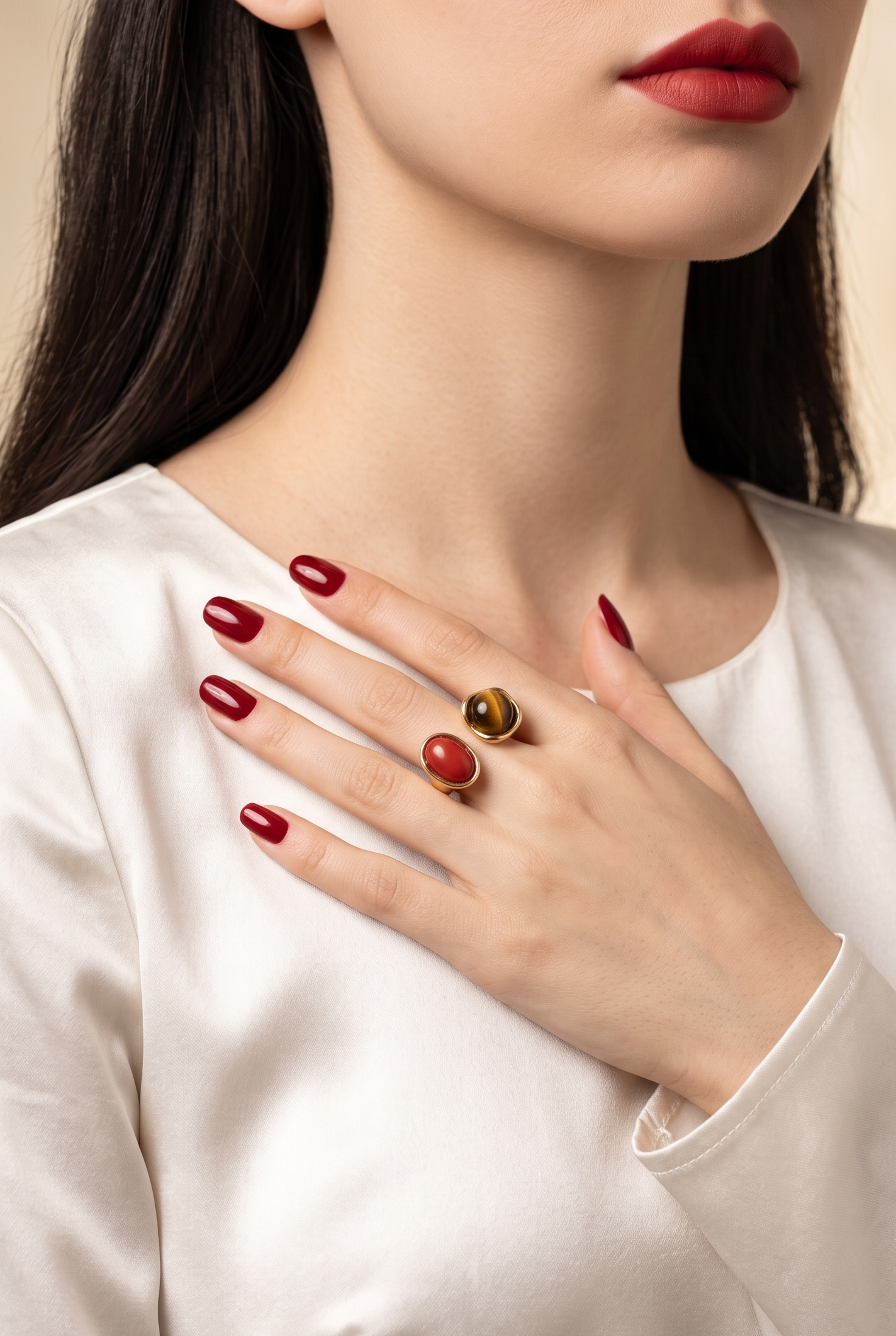 A close-up lifestyle shot of a woman wearing a gold ring with two round red gemstones, styled with red nail polish and a white satin top.