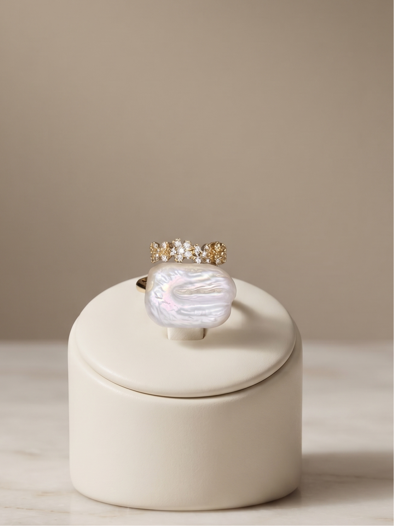 A gold ring with a large iridescent mother-of-pearl cabochon and small clear stones on the band, displayed on a cream-colored cylindrical stand on a marble surface against a warm beige background.