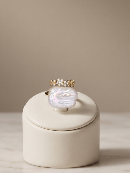 A gold ring with a large iridescent mother-of-pearl cabochon and small clear stones on the band, displayed on a cream-colored cylindrical stand on a marble surface against a warm beige background.