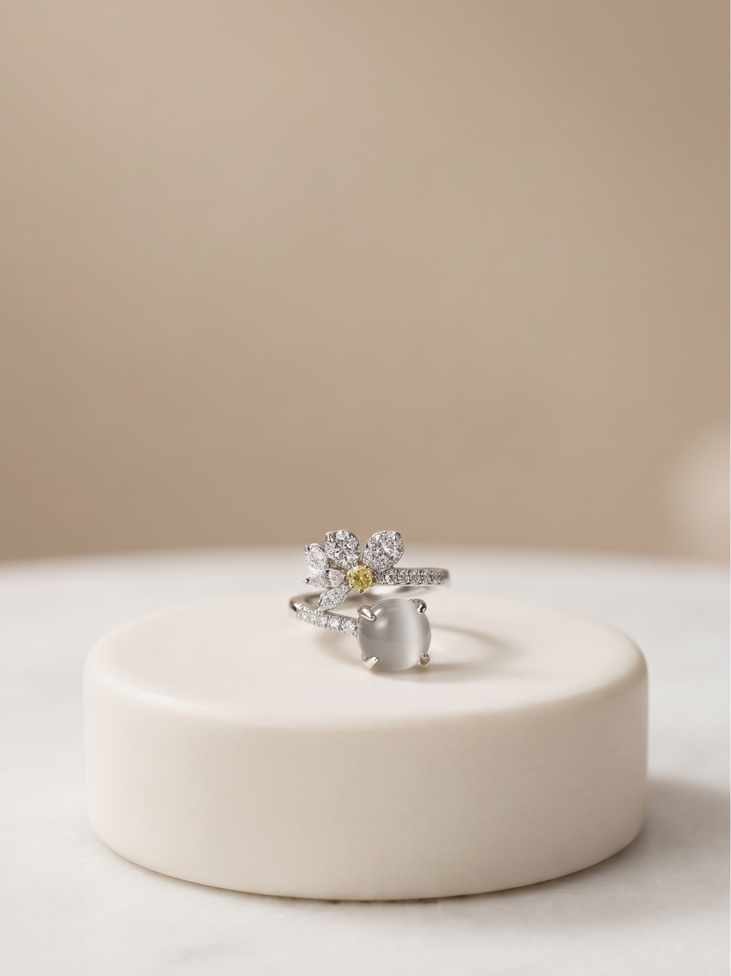 A silver-toned flower ring with a small yellow center stone, displayed on a round cream-colored pedestal against a soft beige background.