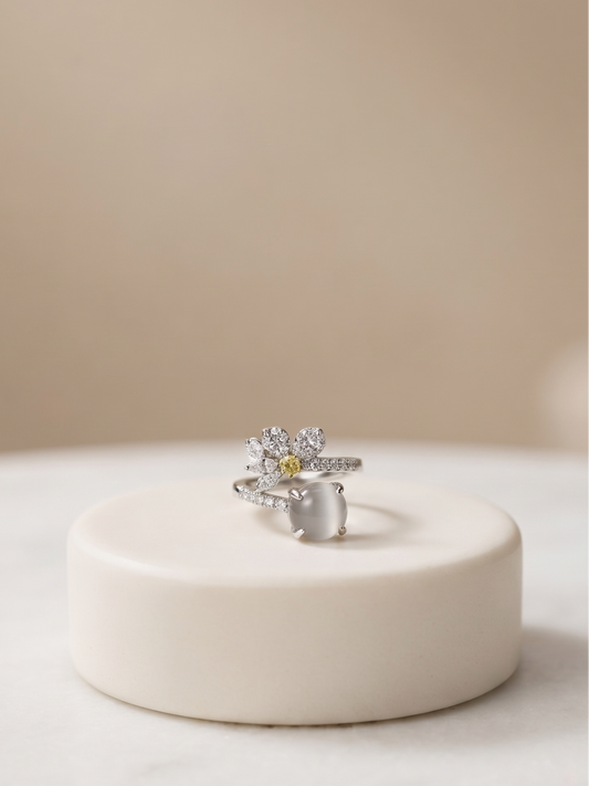 A silver-toned flower ring with a small yellow center stone, displayed on a round cream-colored pedestal against a soft beige background.