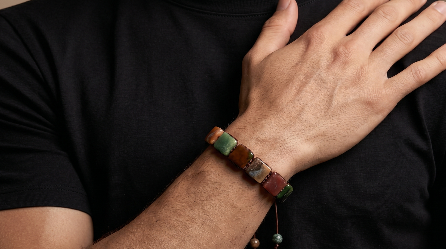 A close-up of a person wearing a beaded bracelet on their wrist, with their hand resting on their chest. The bracelet features a mix of earthy brown, green, and red beads.