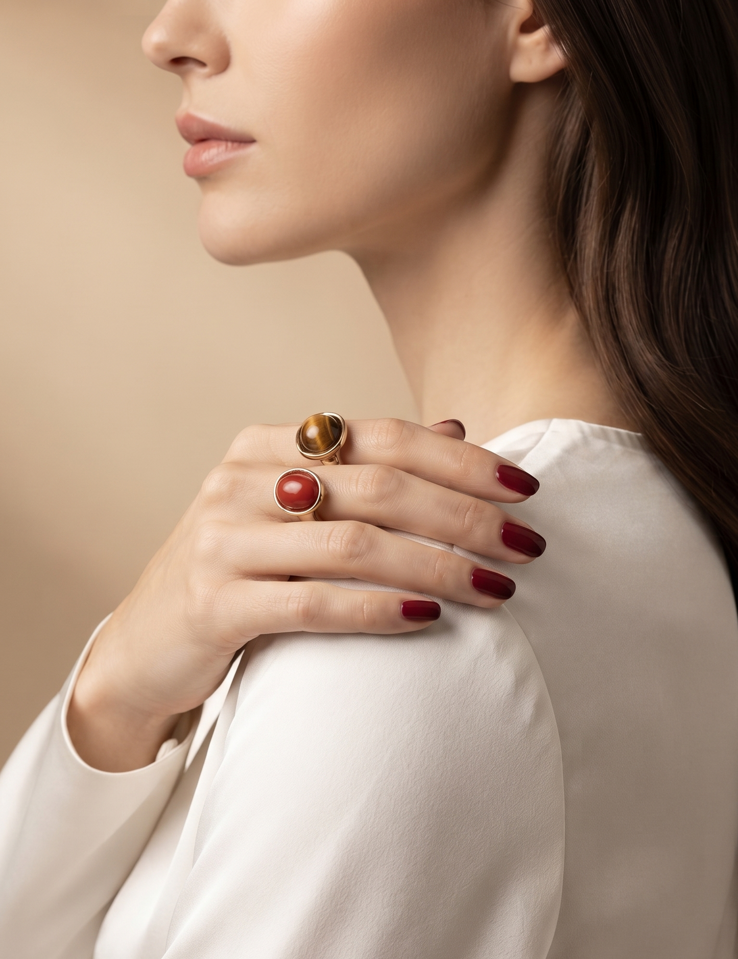 A close-up lifestyle shot of a woman wearing three gold rings on her hand, featuring a large round red stone ring and two smaller tiger's eye rings, paired with a white blouse against a warm beige background.