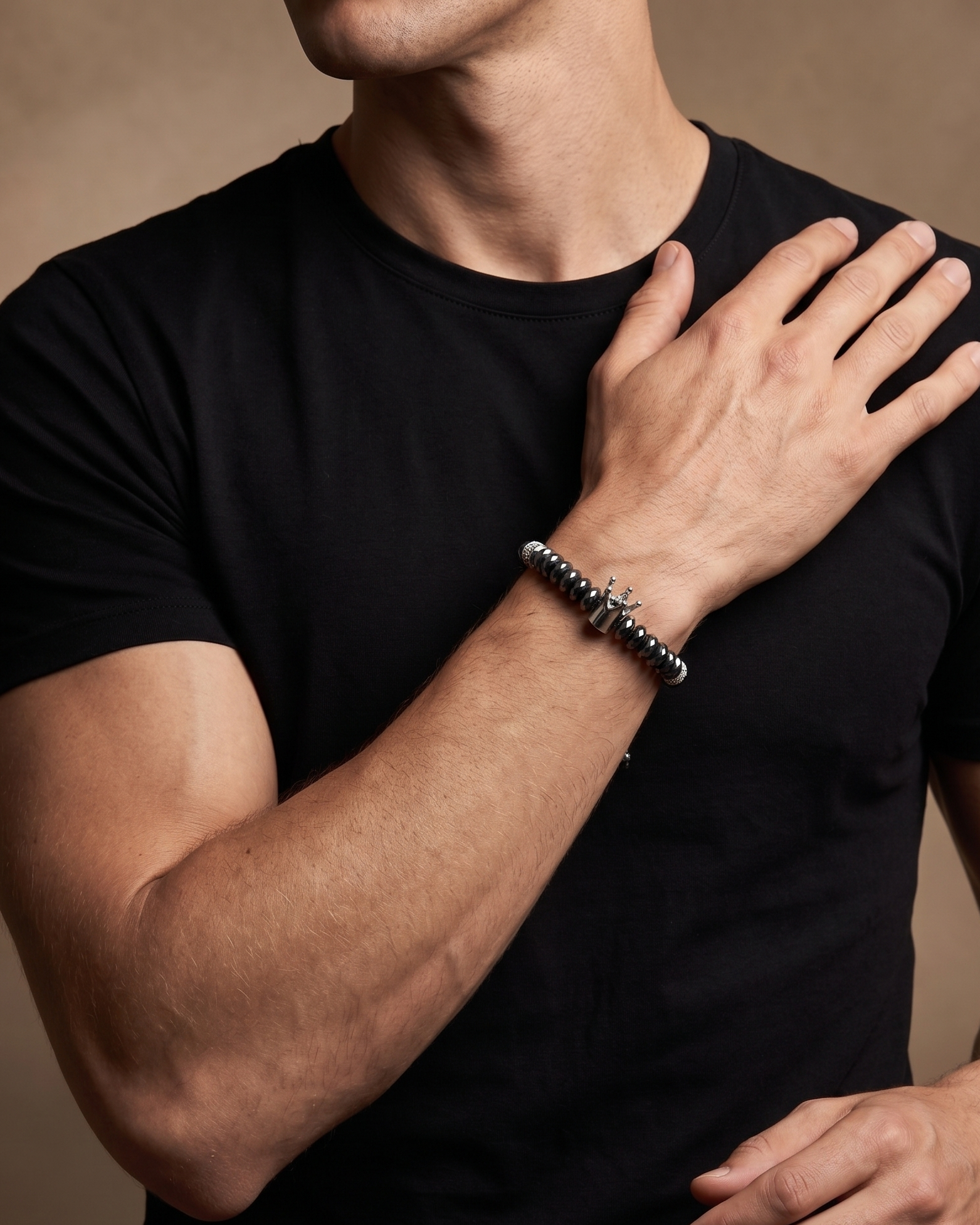 A close-up of a man wearing a black t-shirt and a silver beaded bracelet on his wrist, with his hand resting on his chest against a warm brown background.