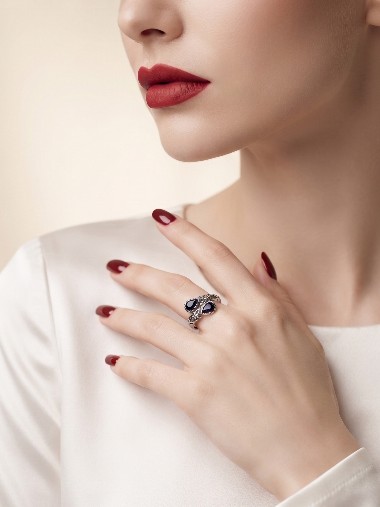A close-up lifestyle shot of a model wearing a silver-toned ring with dark blue oval gemstones, styled with deep red nail polish and a white satin top.