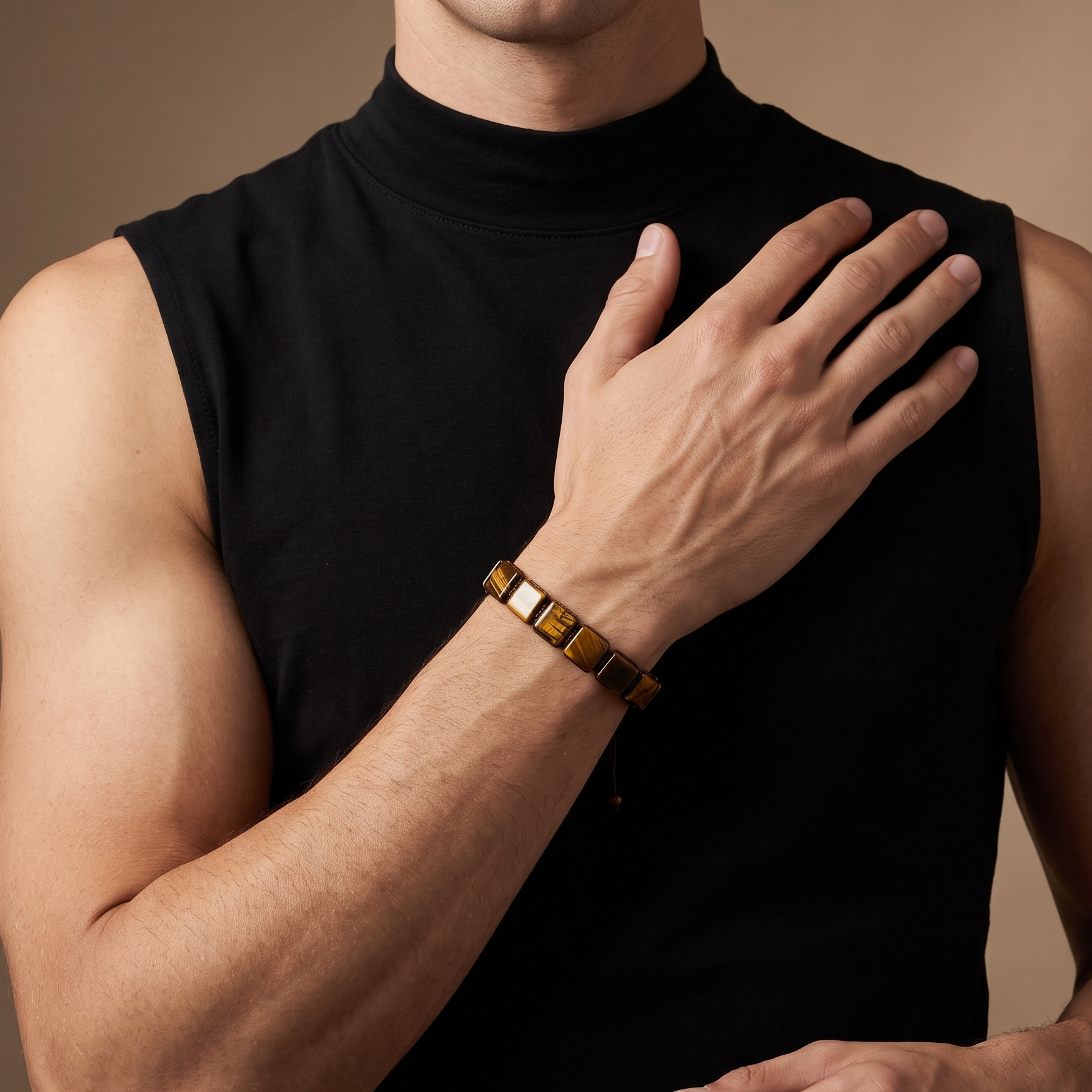 A close-up of a man wearing a black sleeveless mock-neck top and a gold-toned geometric link bracelet on his wrist, set against a warm beige studio background.