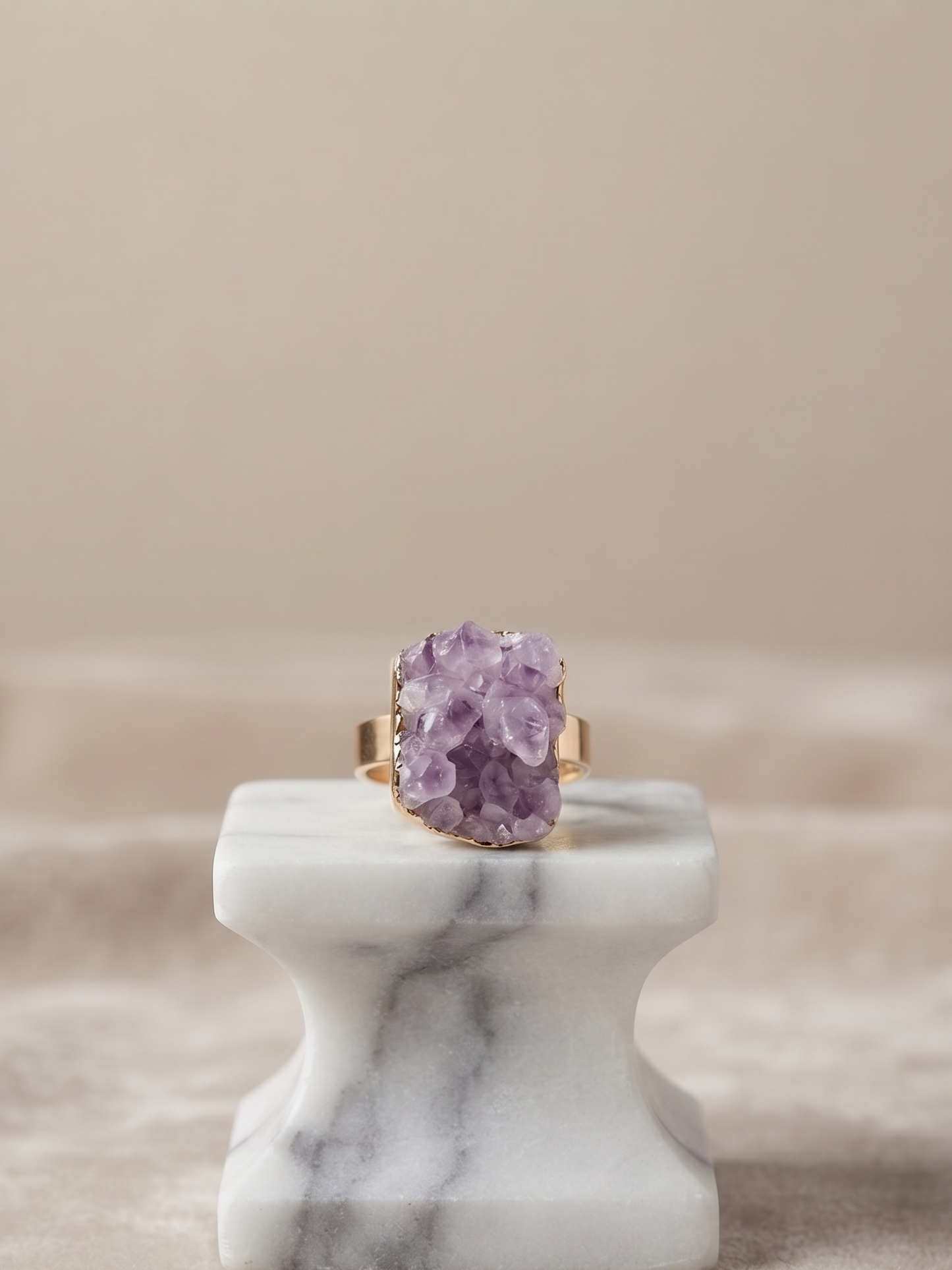 A gold ring with a large, rough-cut purple amethyst crystal cluster, displayed on a white marble ring stand against a soft beige background.