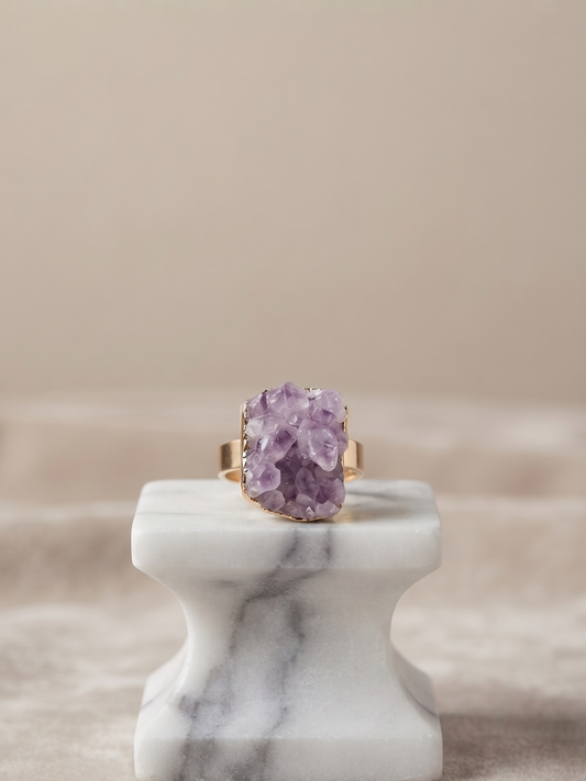 A gold ring with a large, rough-cut purple amethyst crystal cluster, displayed on a white marble ring stand against a soft beige background.