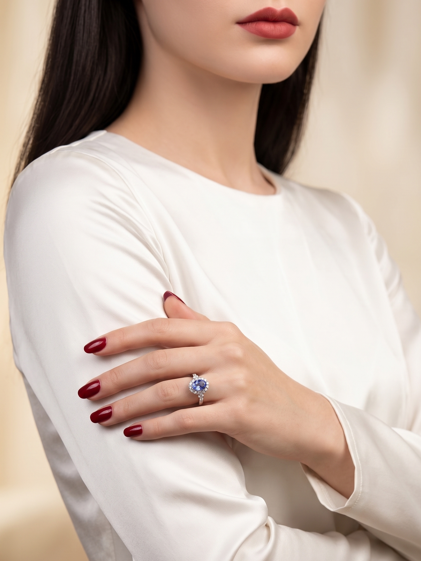 A close-up of a woman wearing a white long-sleeve top, showcasing a ring with a round blue gemstone on her finger. She has dark hair and dark red manicured nails, set against a soft, warm beige background.