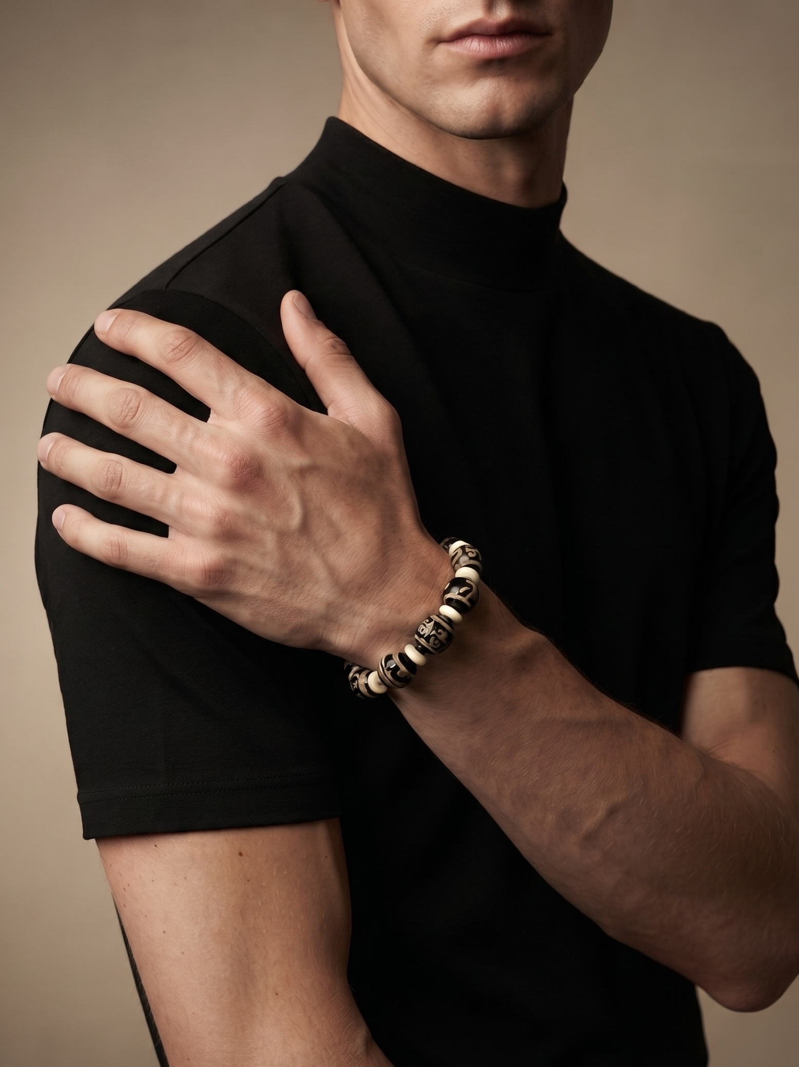 A close-up of a man wearing a black short-sleeve mock neck shirt and a chunky metallic beaded bracelet on his wrist, posed against a solid warm beige background.