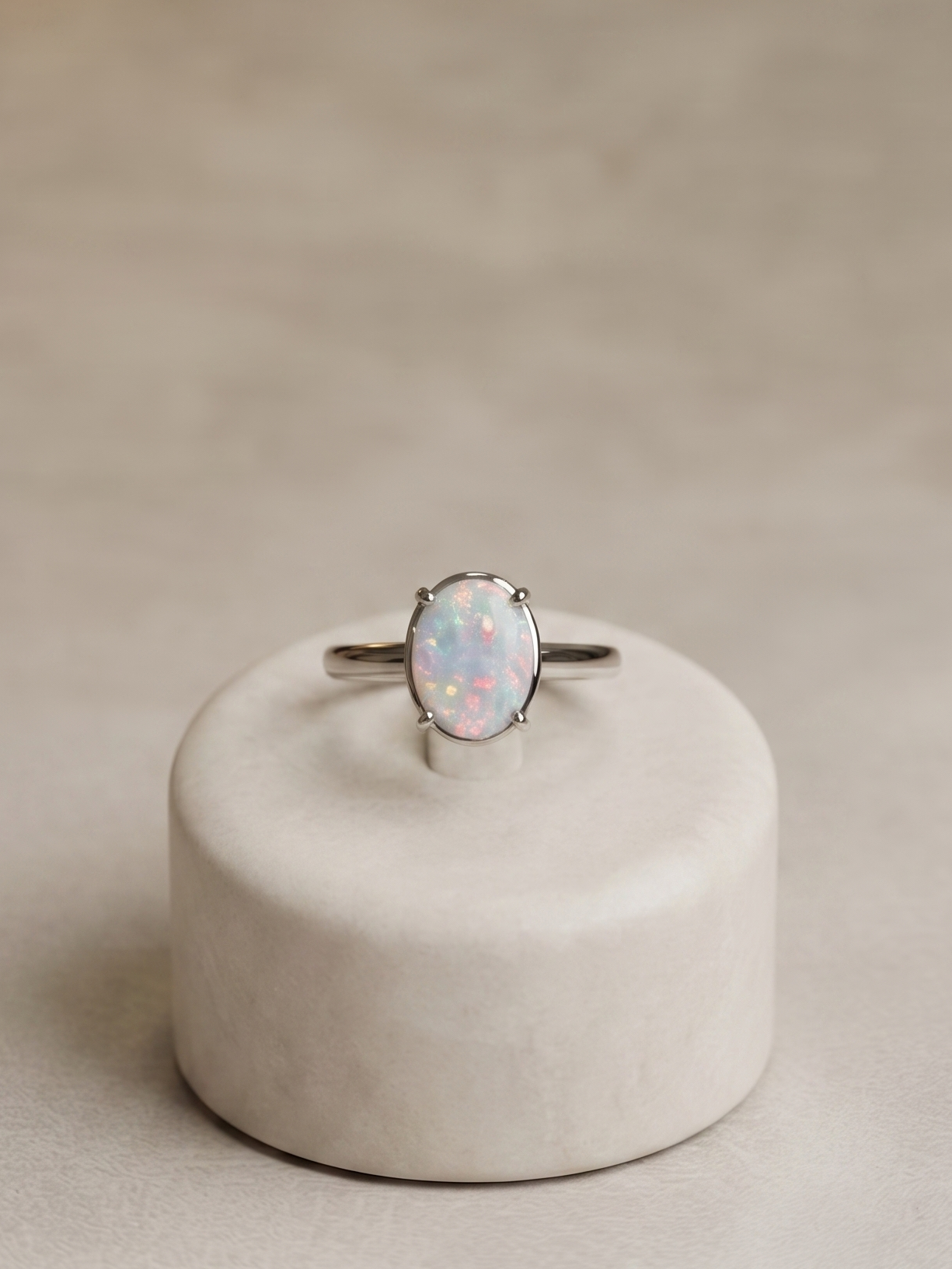 A silver band ring with an oval opal gemstone, displayed on a small round beige pedestal against a neutral beige background.