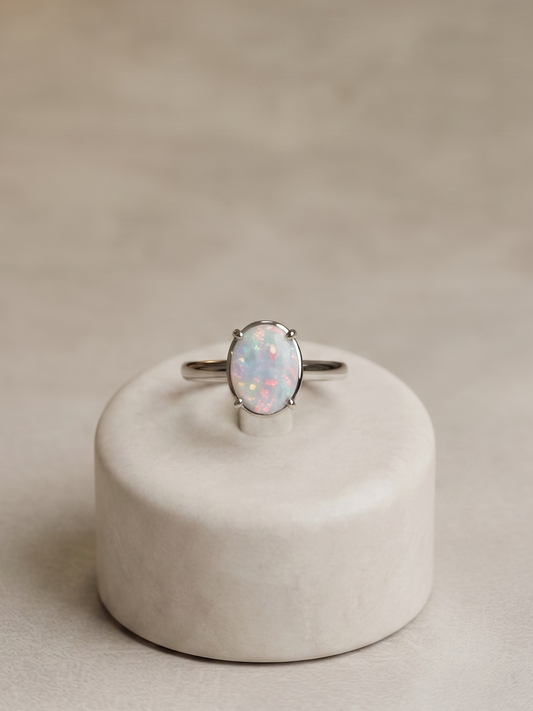 A silver band ring with an oval opal gemstone, displayed on a small round beige pedestal against a neutral beige background.