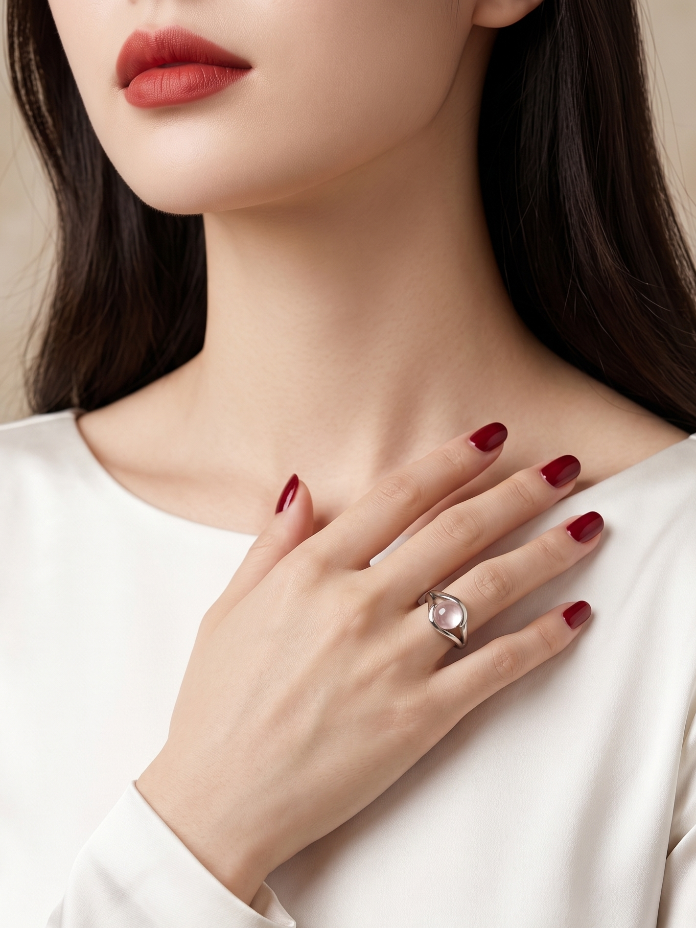 A close-up of a woman wearing a silver ring with a round clear gemstone on her finger, paired with a white long-sleeve top and dark red nail polish.