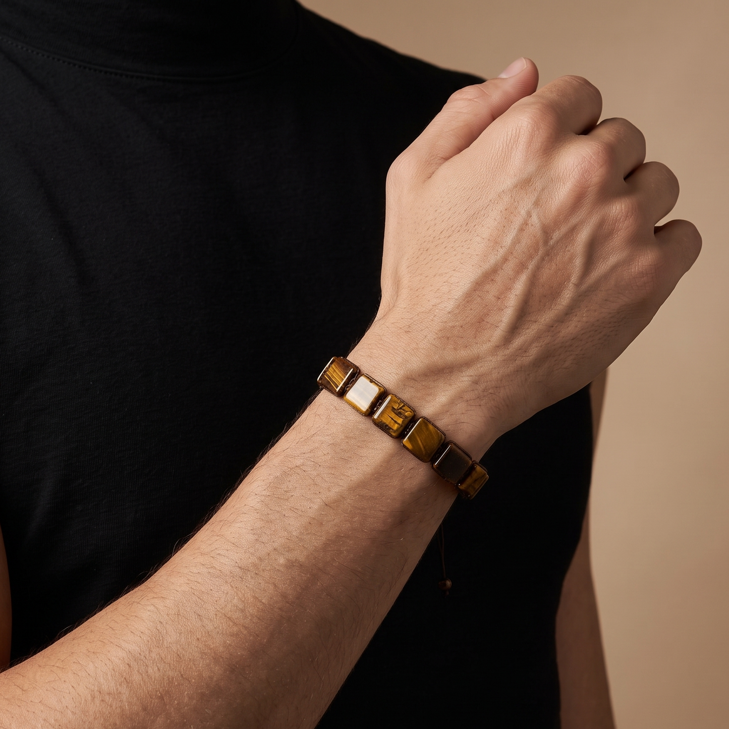 A close-up of a man's arm and hand wearing a gold-toned metal bracelet with alternating polished and matte square links, set against a solid beige background.