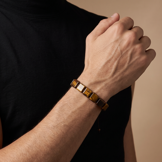 A close-up of a man's arm and hand wearing a gold-toned metal bracelet with alternating polished and matte square links, set against a solid beige background.