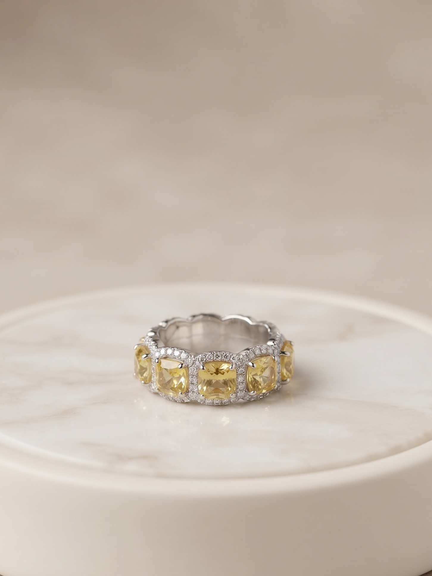 A silver band ring featuring a row of yellow marquise-cut gemstones and small clear accent stones, displayed on a round white marble coaster against a soft beige background.