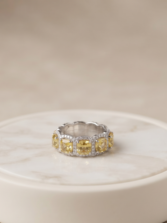 A silver band ring featuring a row of yellow marquise-cut gemstones and small clear accent stones, displayed on a round white marble coaster against a soft beige background.