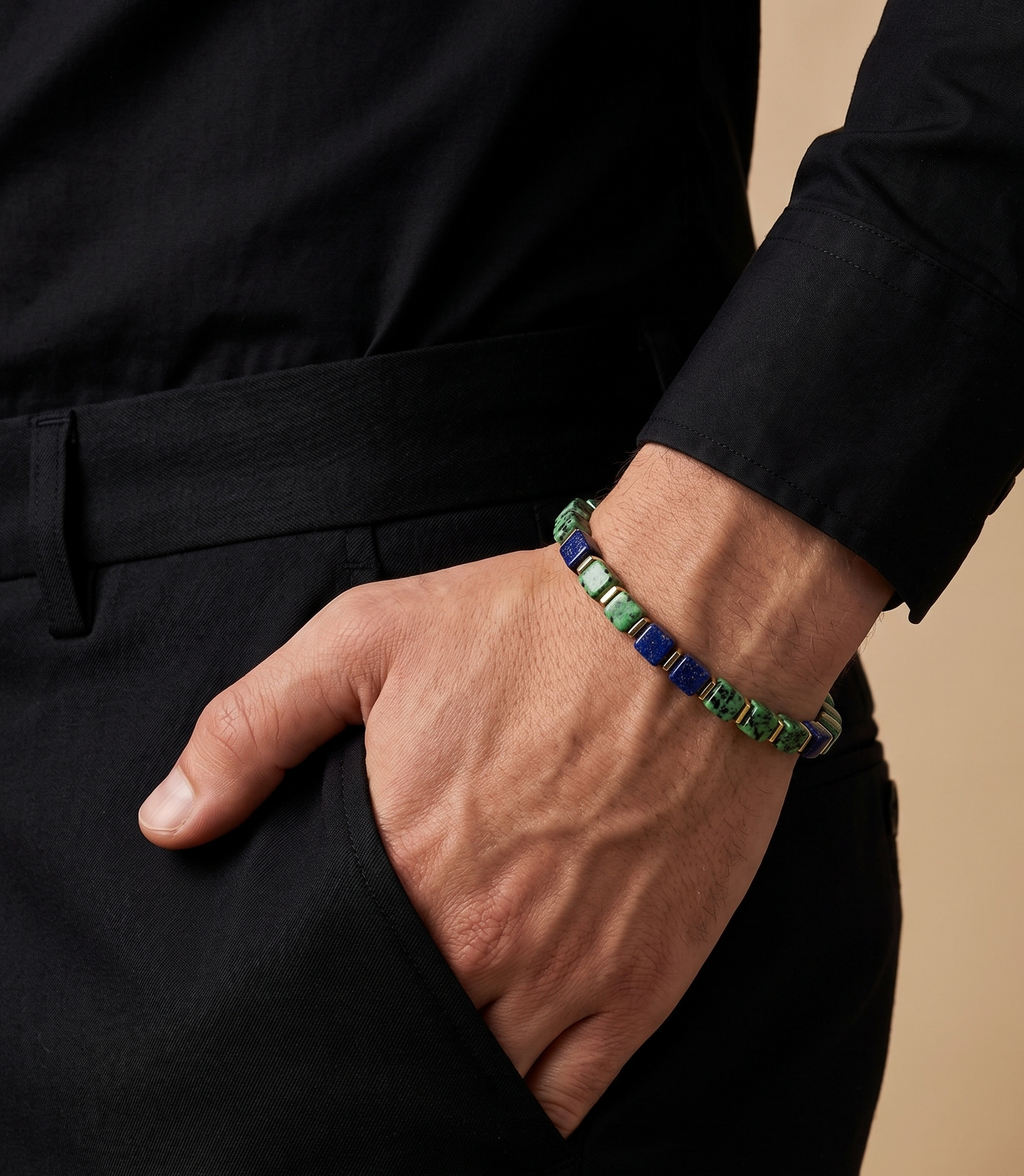 A close-up of a person's hand in a black pocket wearing a beaded bracelet with green, blue, and white beads, paired with a black shirt and trousers against a beige background.