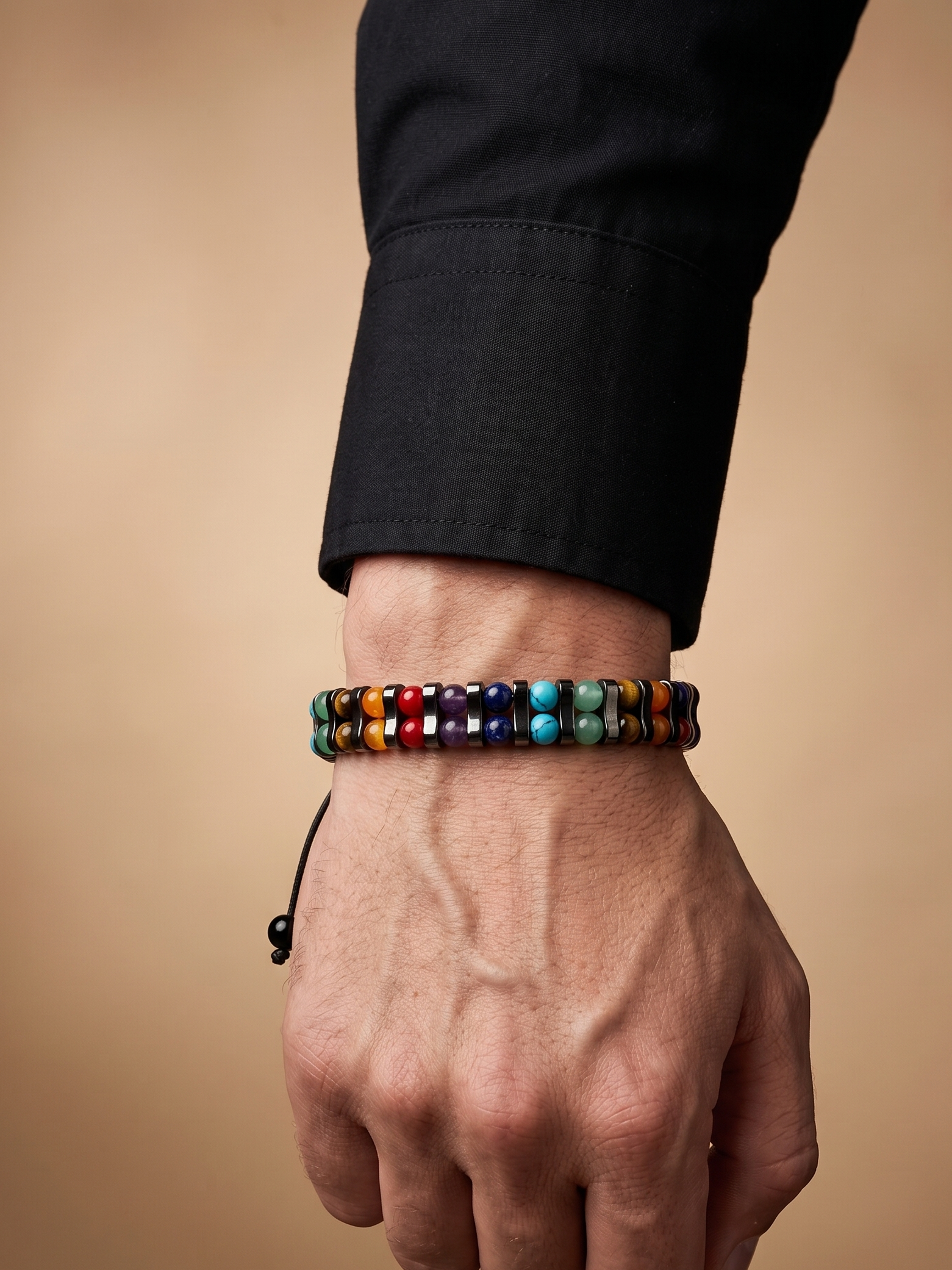A close-up of a person's hand and wrist wearing a beaded bracelet made of small multicolored round beads, with a black cuff sleeve visible on the arm against a warm beige background.