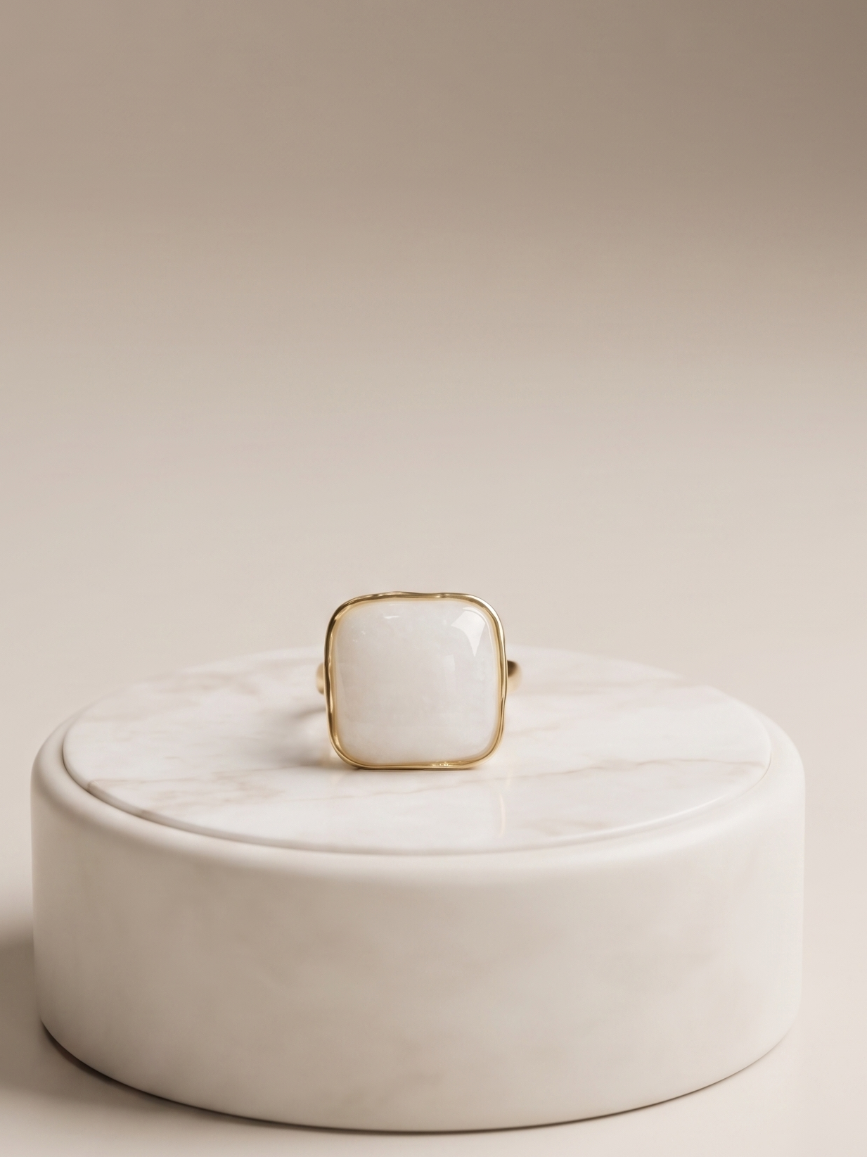 A gold ring with a square white cabochon gemstone, displayed on top of a round white marble box against a warm beige background.
