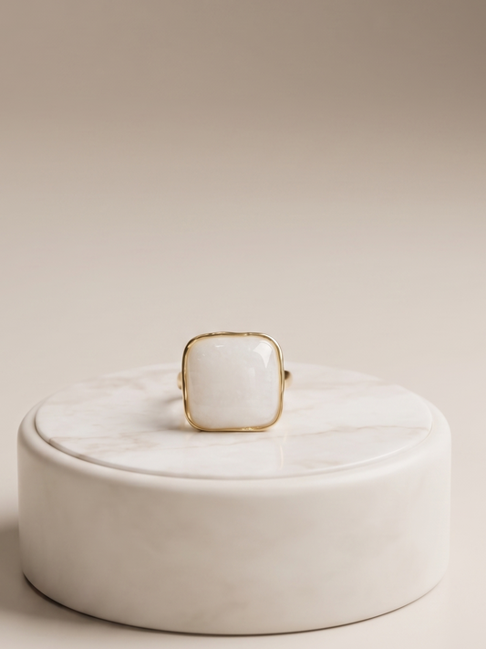A gold ring with a square white cabochon gemstone, displayed on top of a round white marble box against a warm beige background.