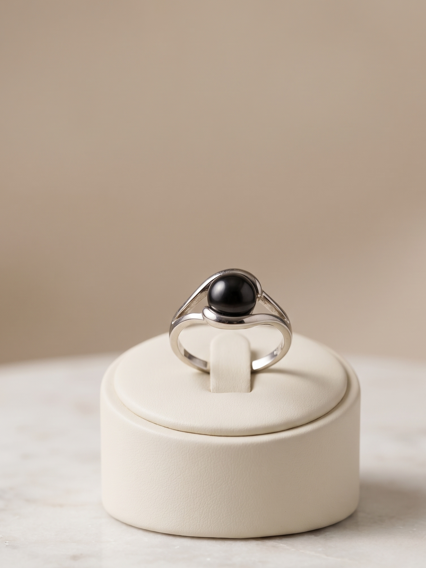 A silver ring with a large black oval stone, displayed on a cream-colored jewelry stand on a light marble surface against a warm beige background.