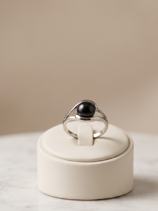 A silver ring with a large black oval stone, displayed on a cream-colored jewelry stand on a light marble surface against a warm beige background.