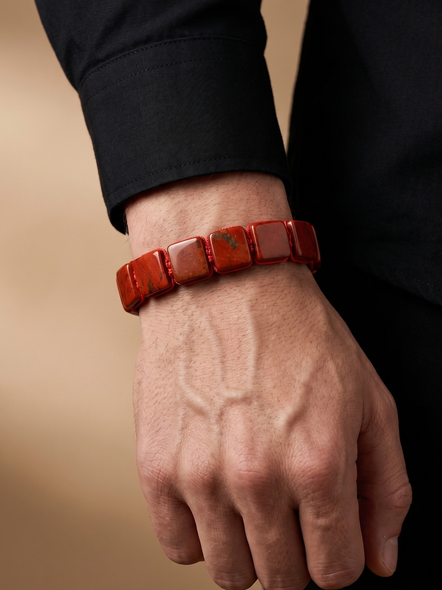 A close-up of a person's hand wearing a red beaded bracelet made of square polished stone tiles, set against a dark shirt and a soft beige background.