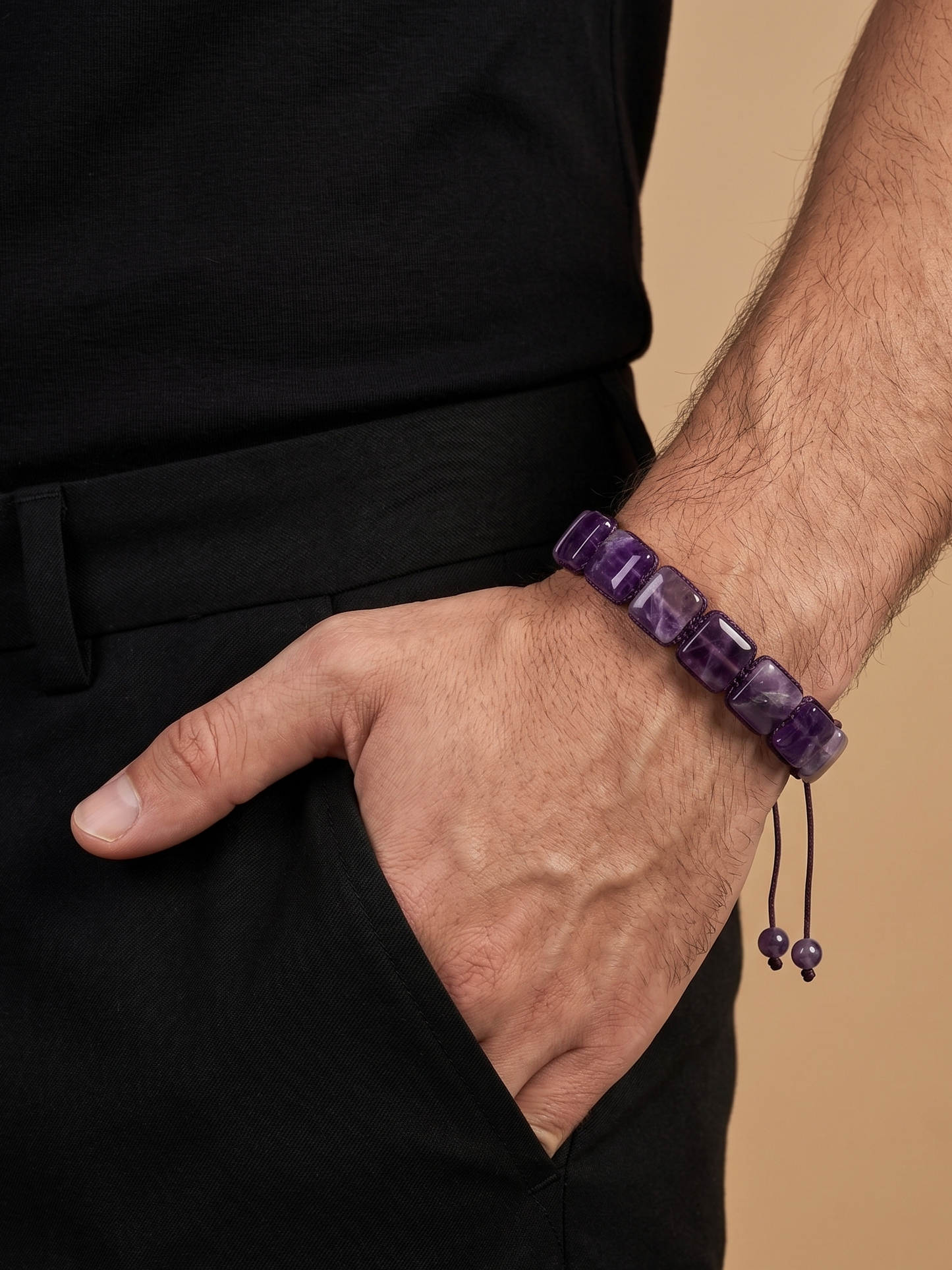 A close-up of a man's wrist wearing a purple beaded bracelet with dark cord and bead tassels, paired with a black shirt and trousers against a warm beige background.