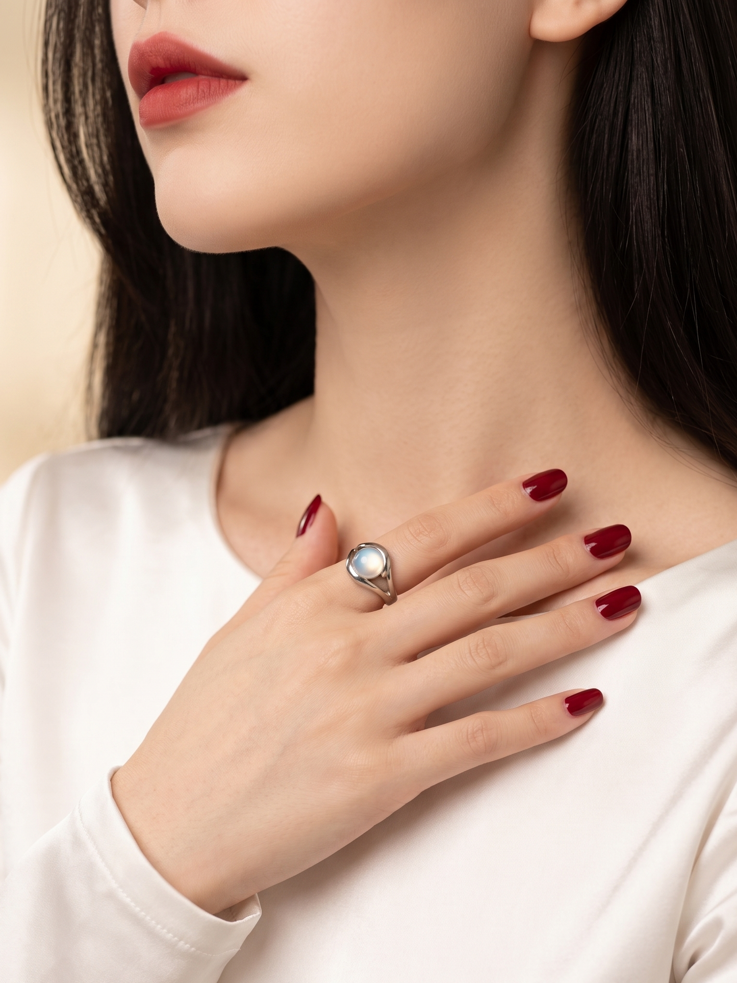 A close-up lifestyle shot of a woman wearing a silver ring with a round clear gemstone on her finger, paired with a white long-sleeve top and dark red manicured nails.
