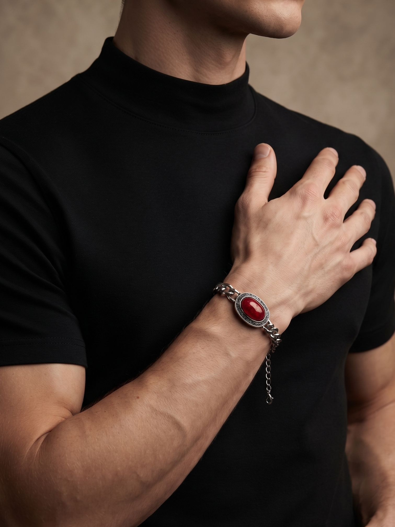 A close-up of a man wearing a black turtleneck shirt and a silver-toned chain bracelet with a prominent oval red cabochon stone, set against a textured warm brown background.