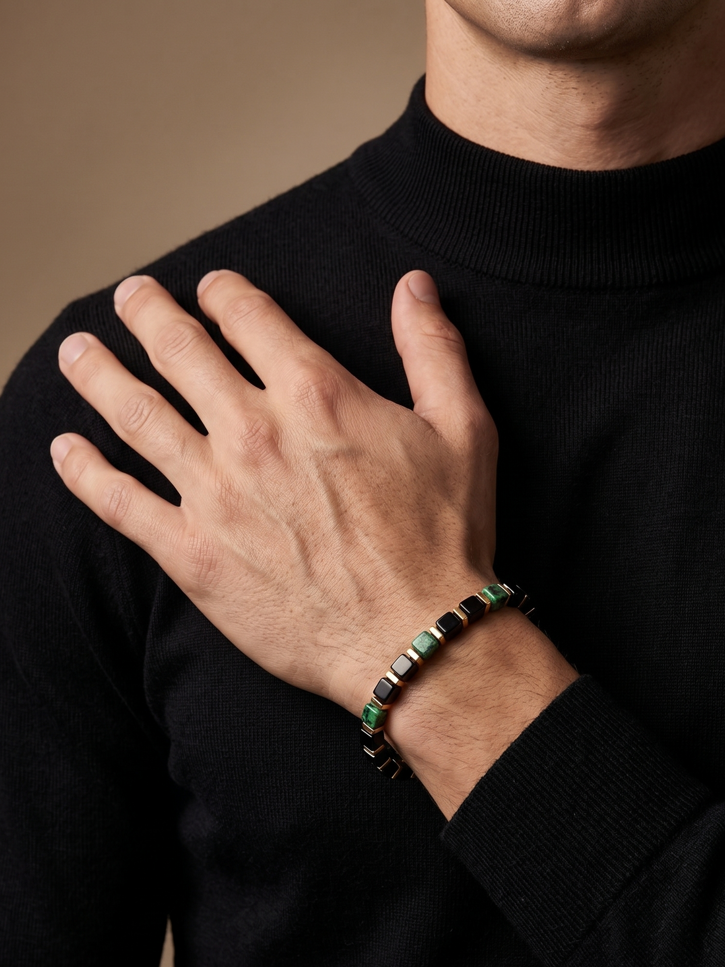 A close-up of a man wearing a black turtleneck sweater and a beaded bracelet on his wrist, with his hand resting on his chest against a warm beige background.